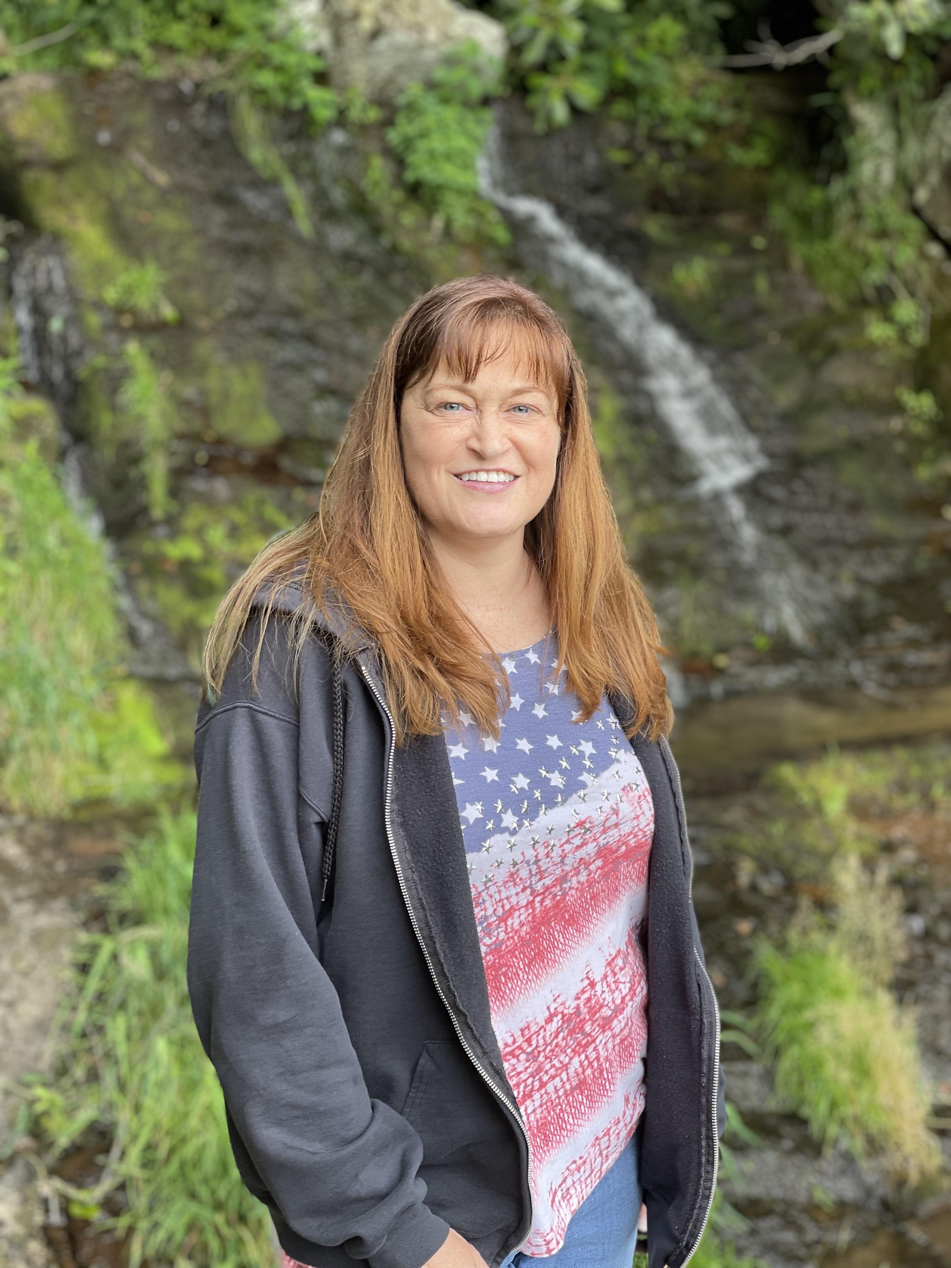 A smiling woman stands beside a serene waterfall, surrounded by vibrant greenery and sunlight.