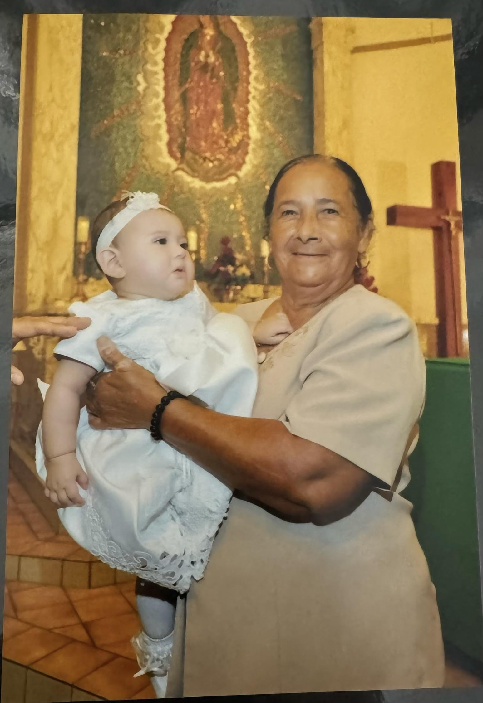 A grandmother joyfully holds her grandchild dressed in white during a baptism in a church.