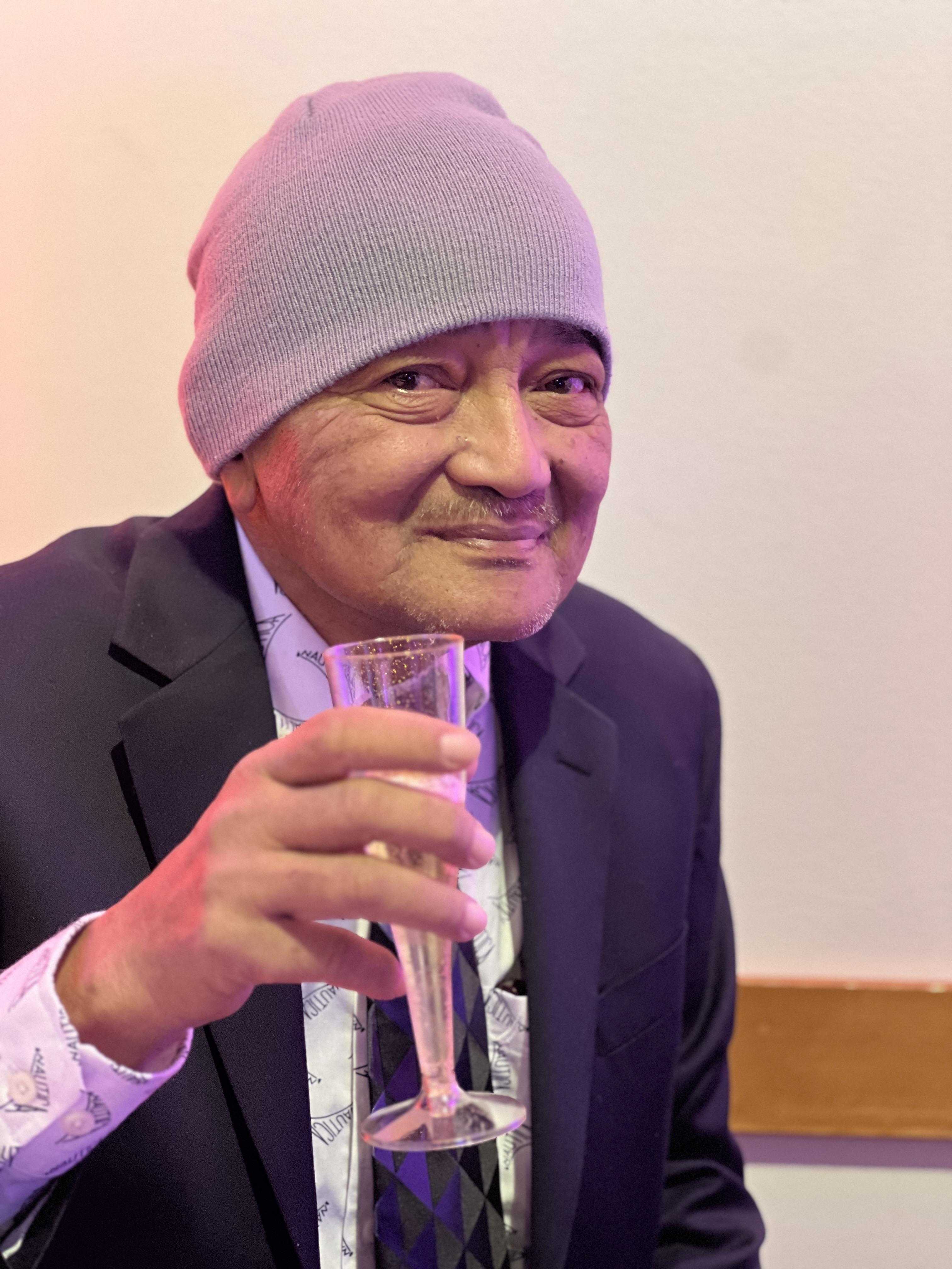 Smiling elder in formal attire enjoys a drink, exuding warmth and joy in a relaxed environment.
