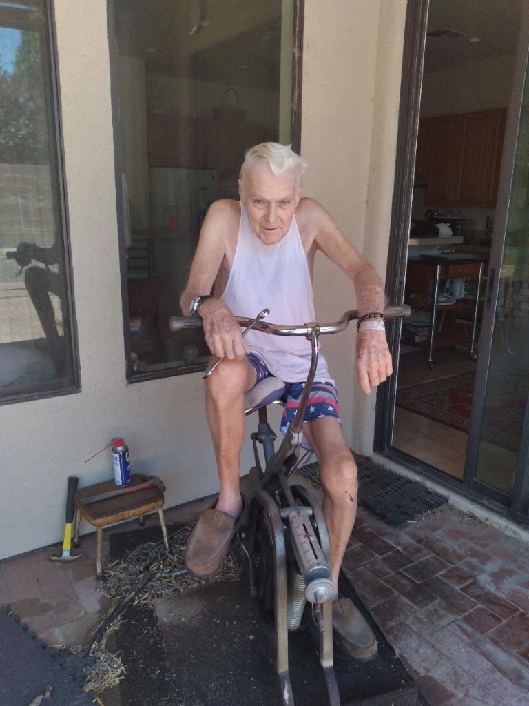 Older man pedals on a stationary bike inside a well-lit room with large windows.