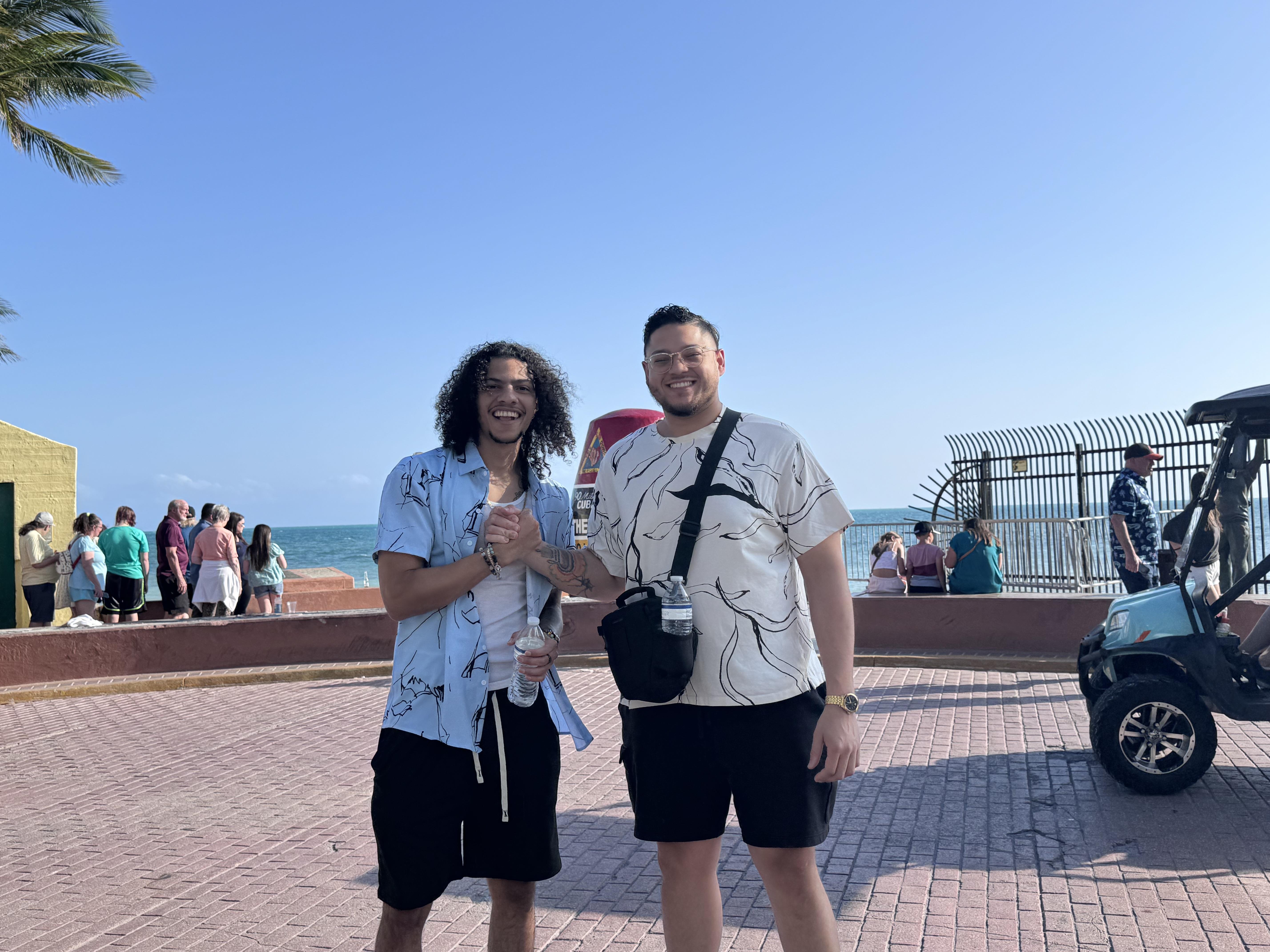 Two men are smiling and posing together at a vibrant beach promenade under bright blue skies.