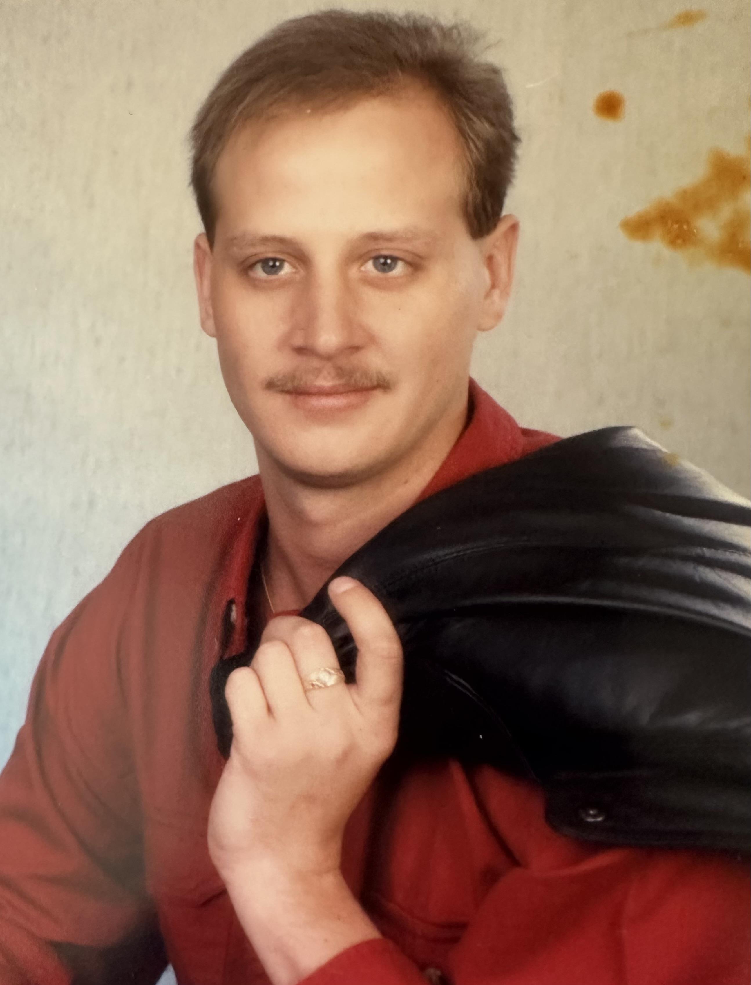 A man wearing a red shirt stands confidently, casually holding a black jacket on his shoulder.