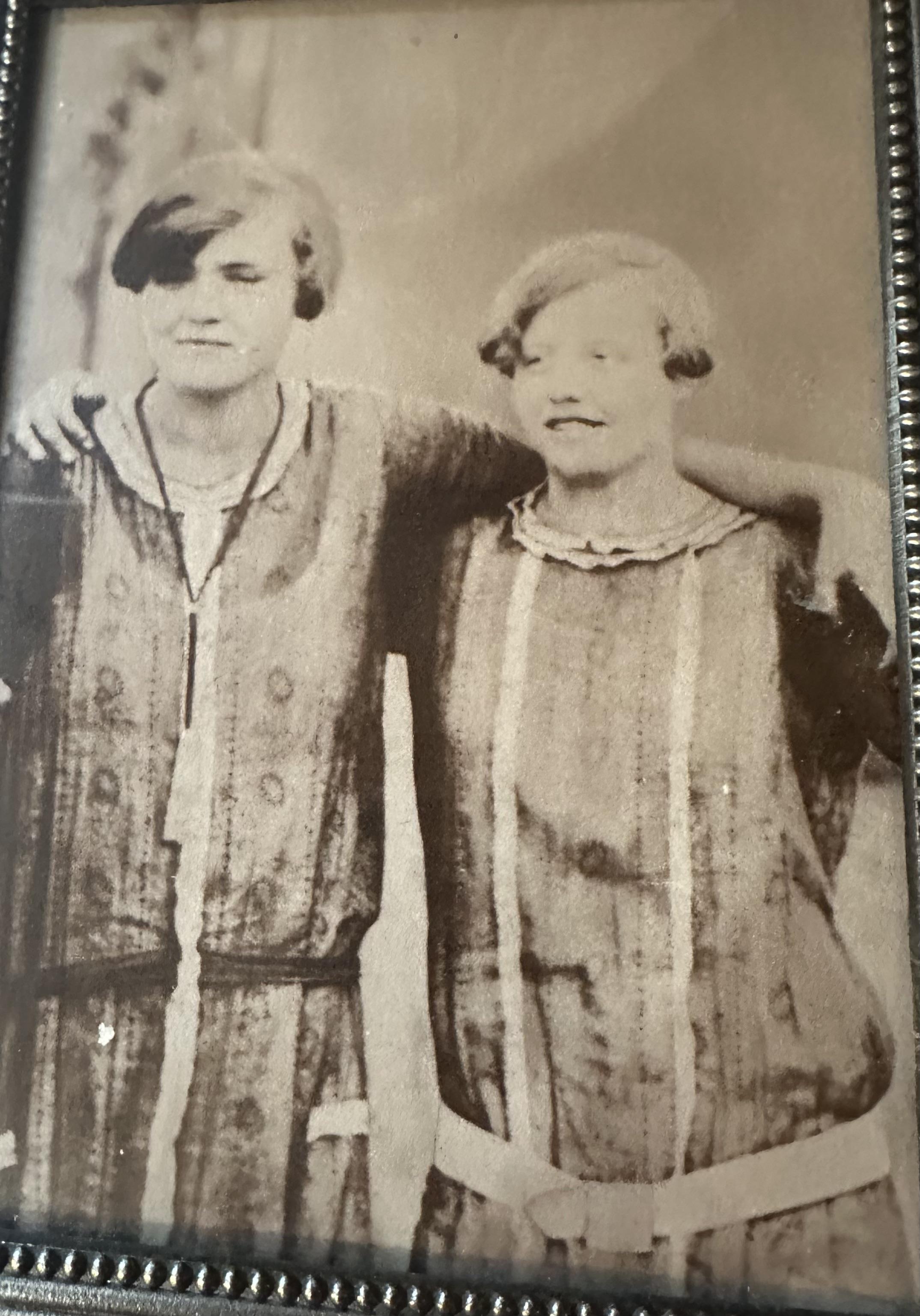 Two women stand side by side, dressed in early 20th century outfits, sharing a joyful moment.