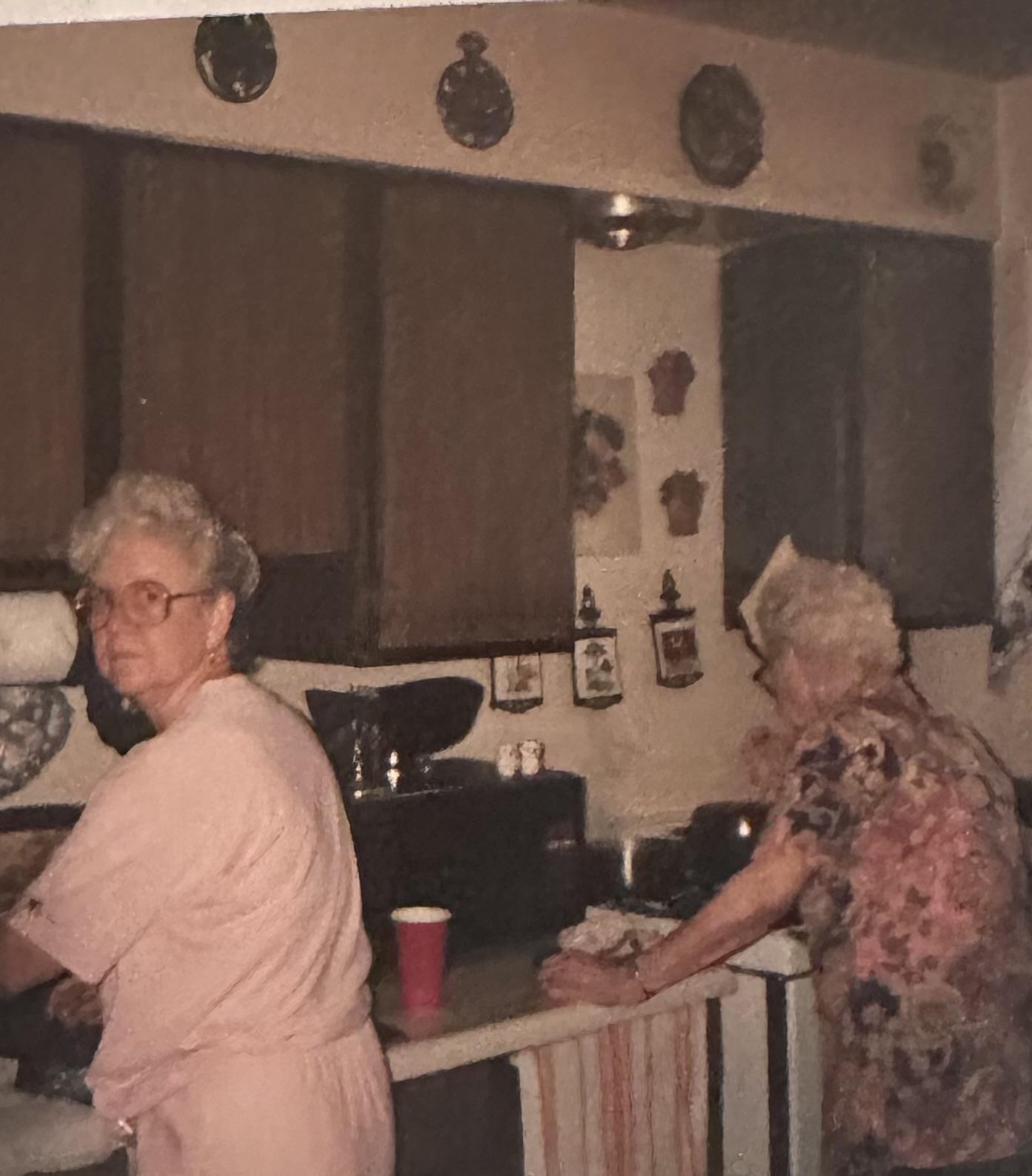 Two elderly women engage in meal preparation in a warm kitchen filled with nostalgic items.