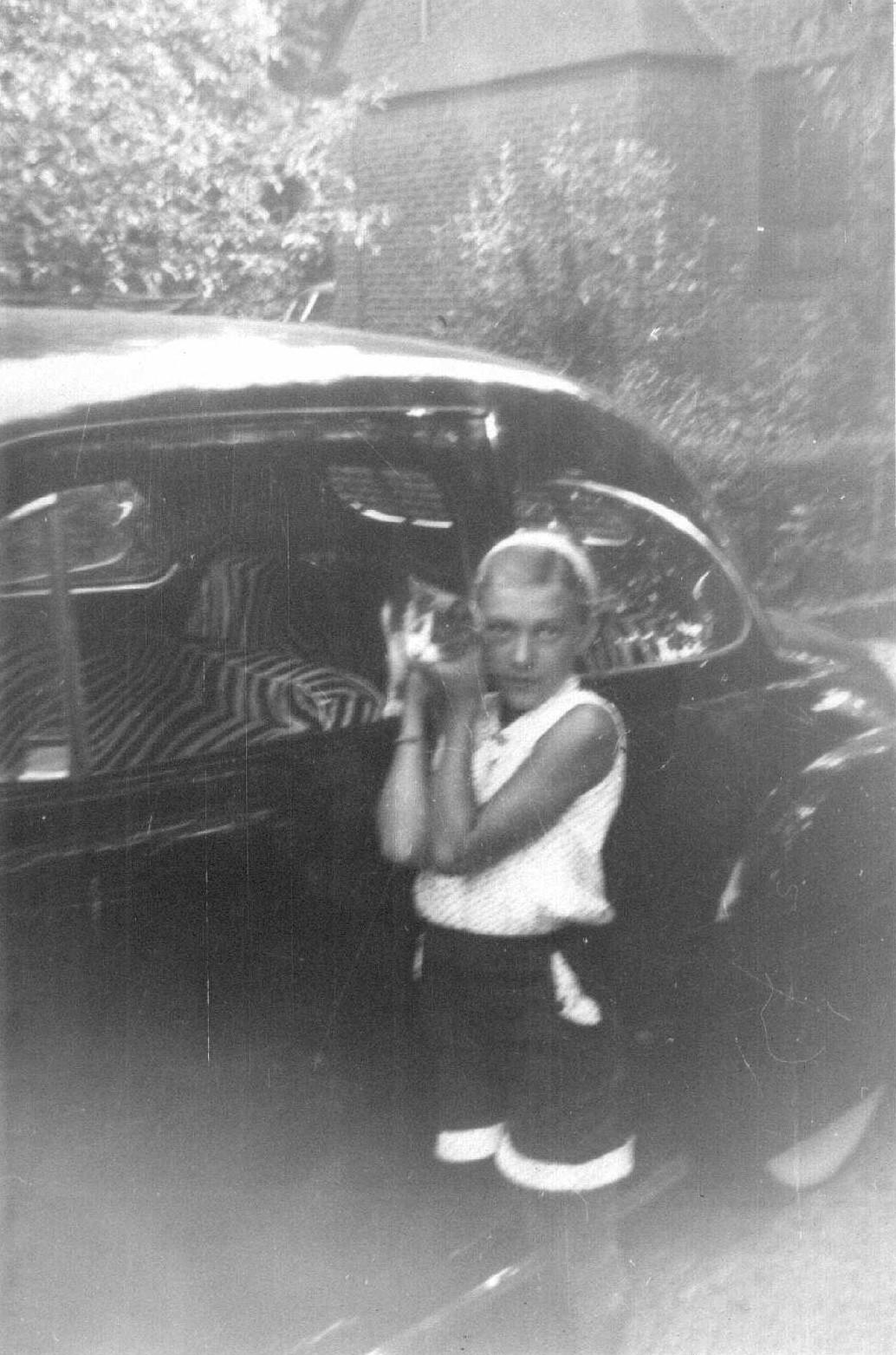 Child stands beside an old car, smiling and holding an object in playful stance on a bright day.