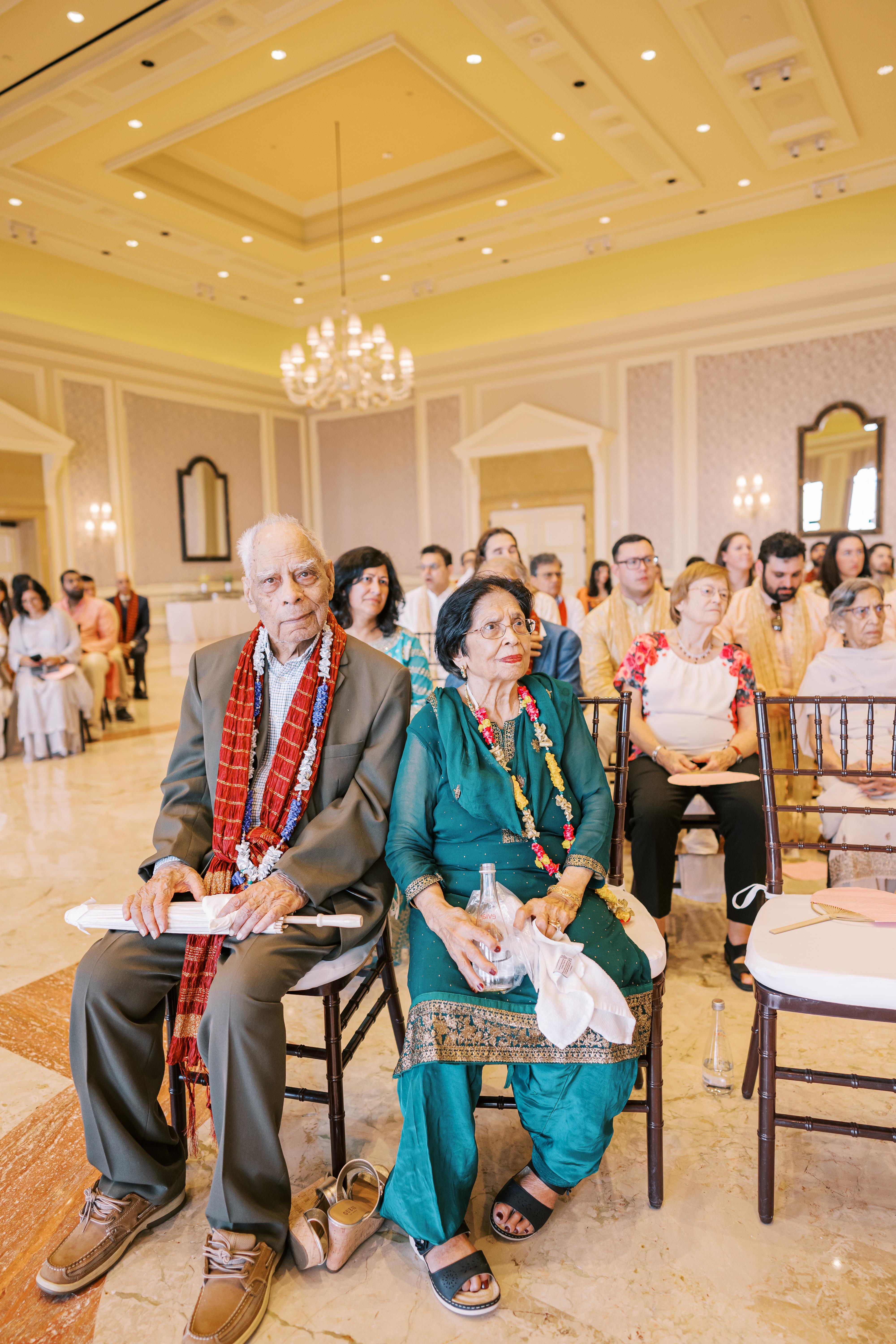 An elderly couple in traditional attire smiles at a ceremonial event with guests nearby.
