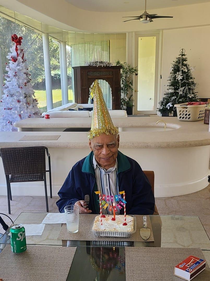 An elderly person sits at a table celebrating a birthday with a decorated cake and festive decor.