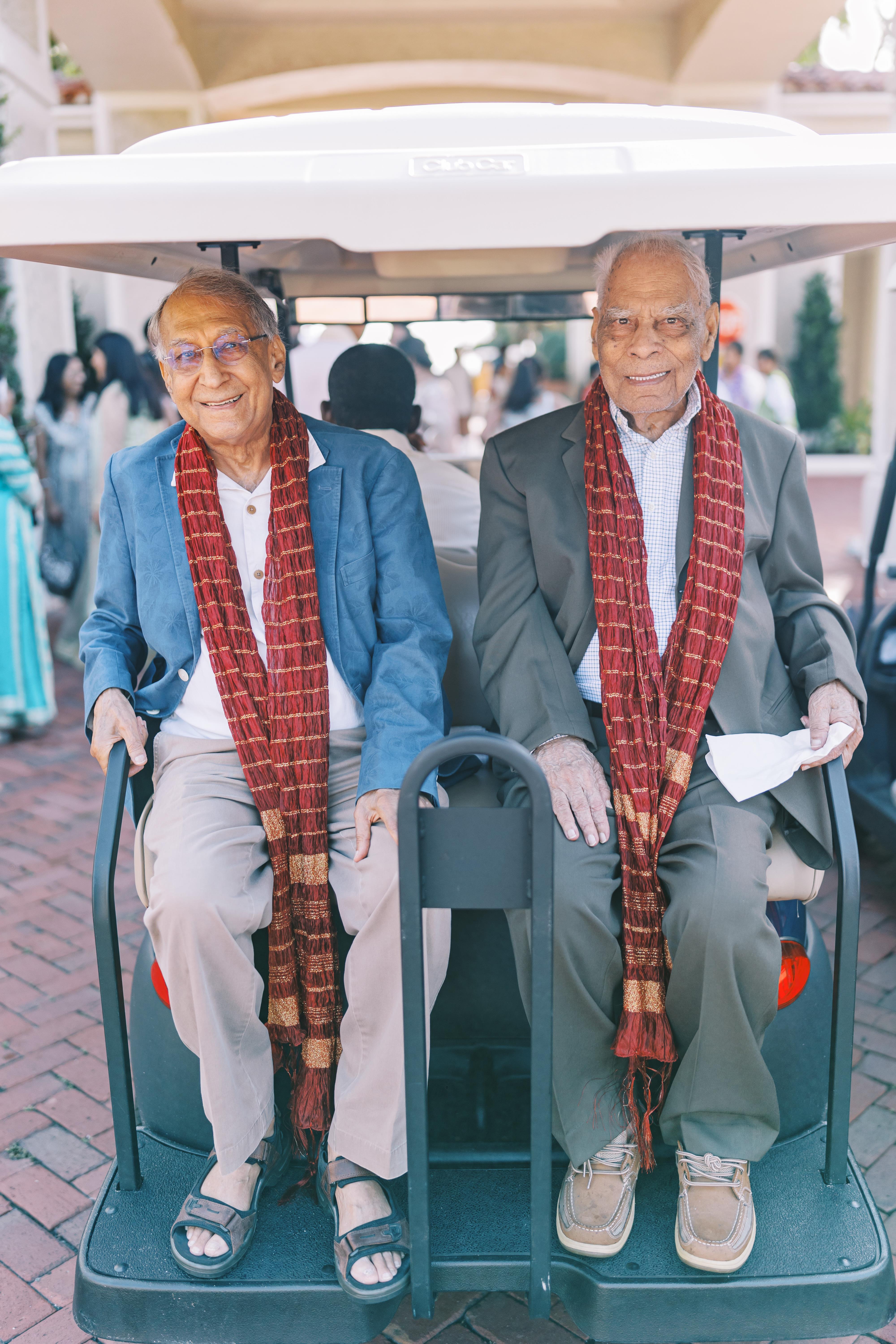 Two smiling elderly men in a golf cart, wearing scarves and enjoying the festivities.
