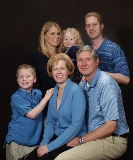A family gathers closely, smiling and wearing coordinated blue outfits, enjoying a moment together.