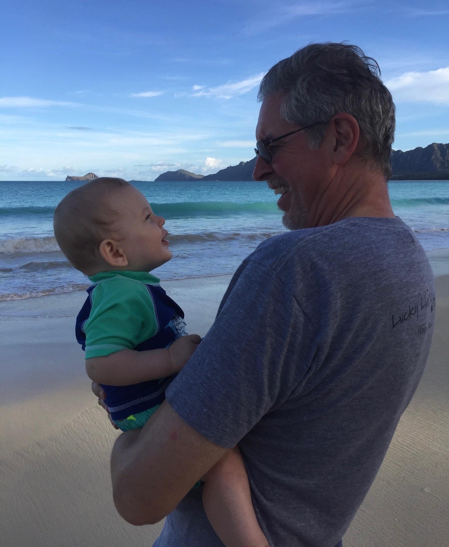 Grandfather holds his smiling grandson by the shoreline, enjoying a vibrant beach day.