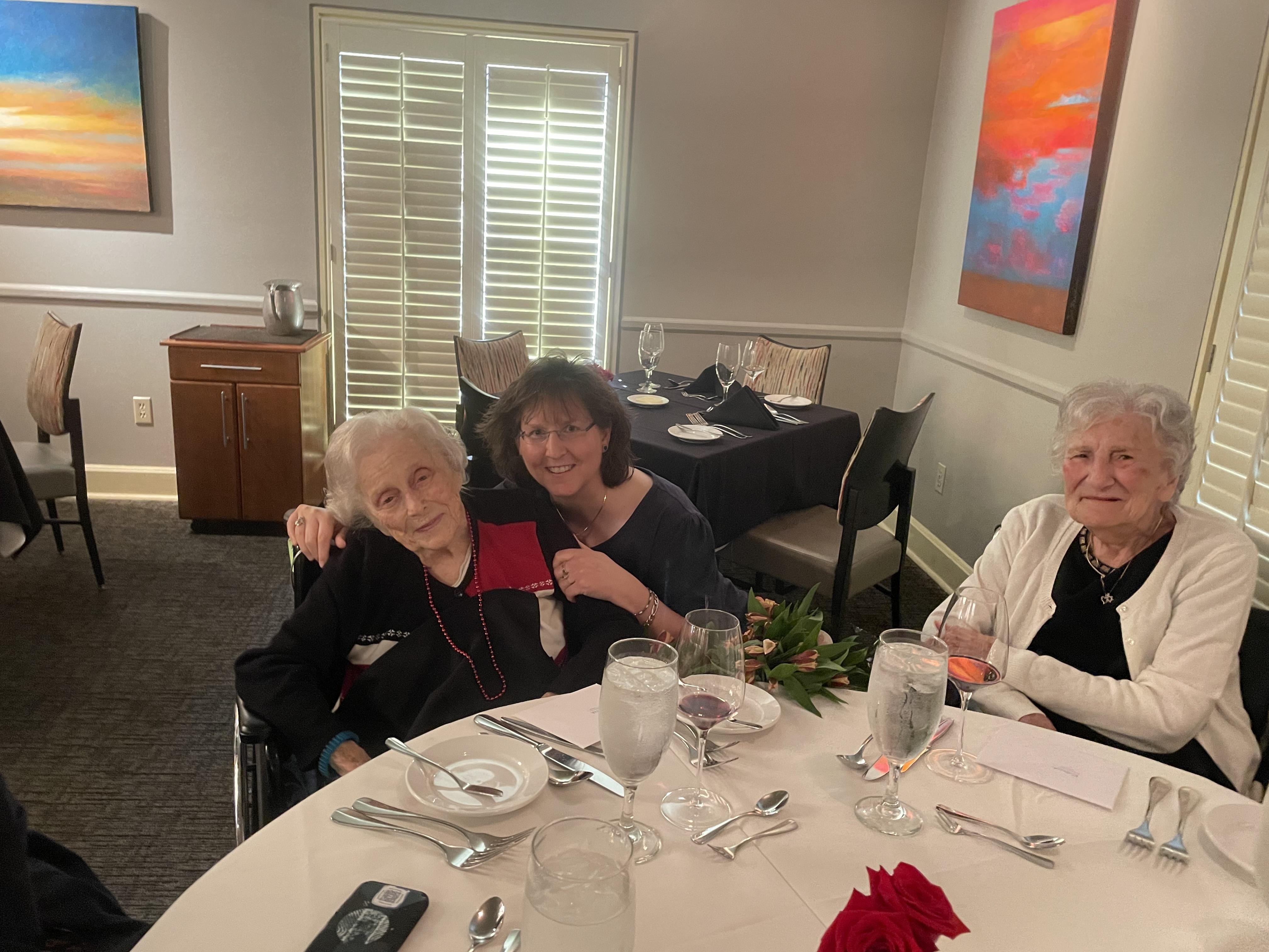 Two women warmly embrace an elderly lady at a beautifully set dining table while smiling.