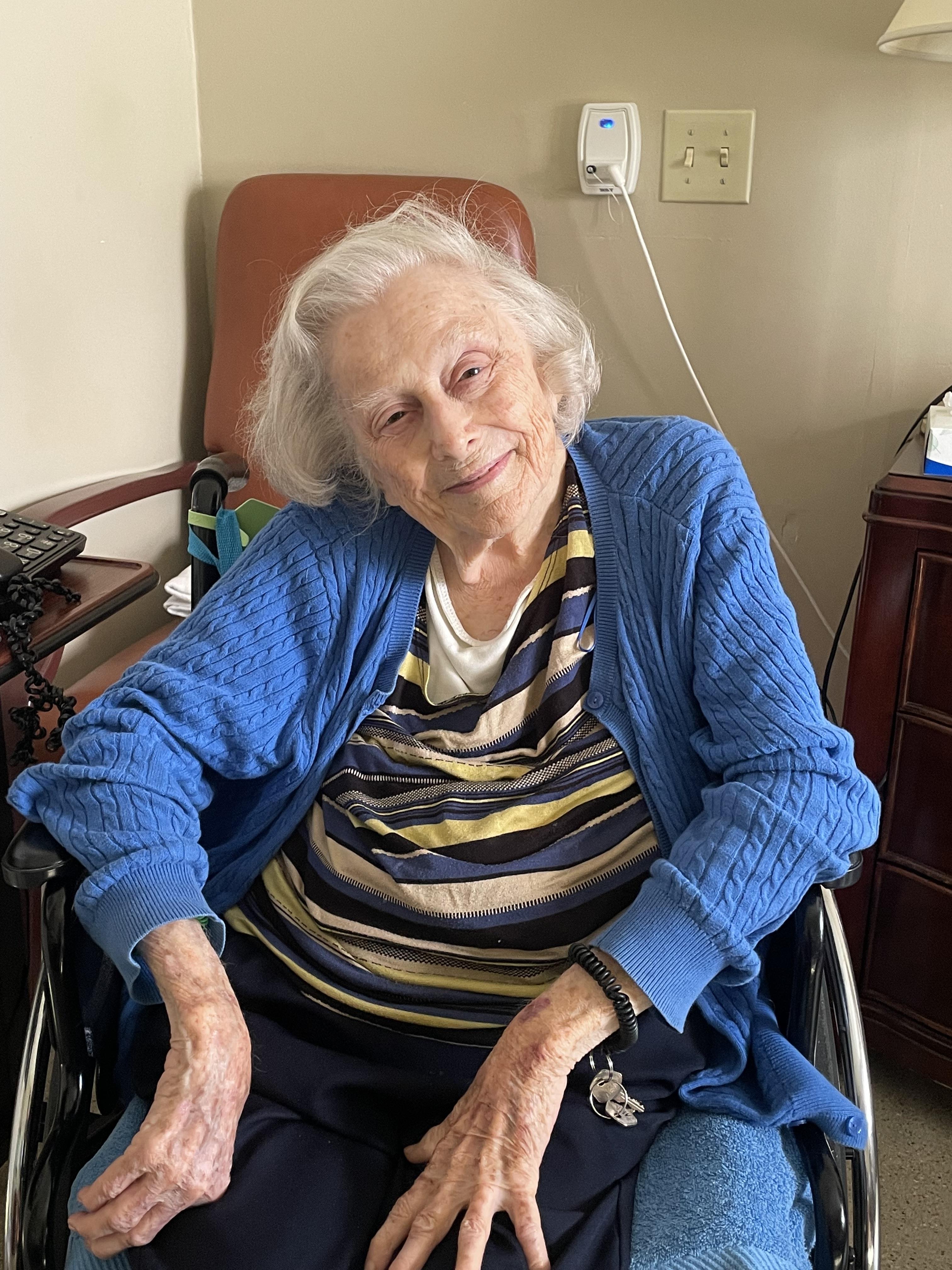 An elderly woman in a striped shirt and blue cardigan smiles while in a wheelchair.