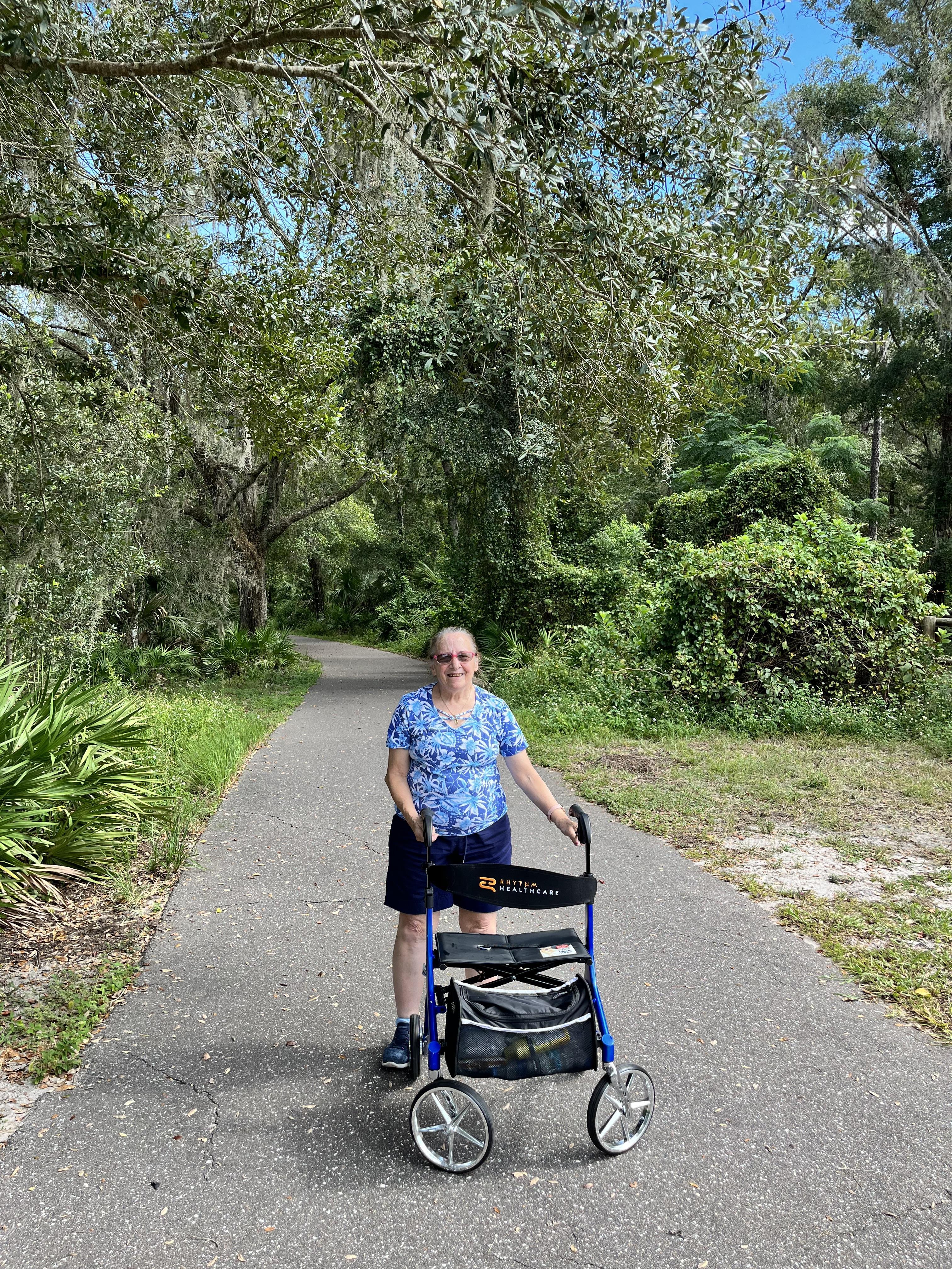 A woman walks confidently along a serene path with a walker, surrounded by lush greenery.