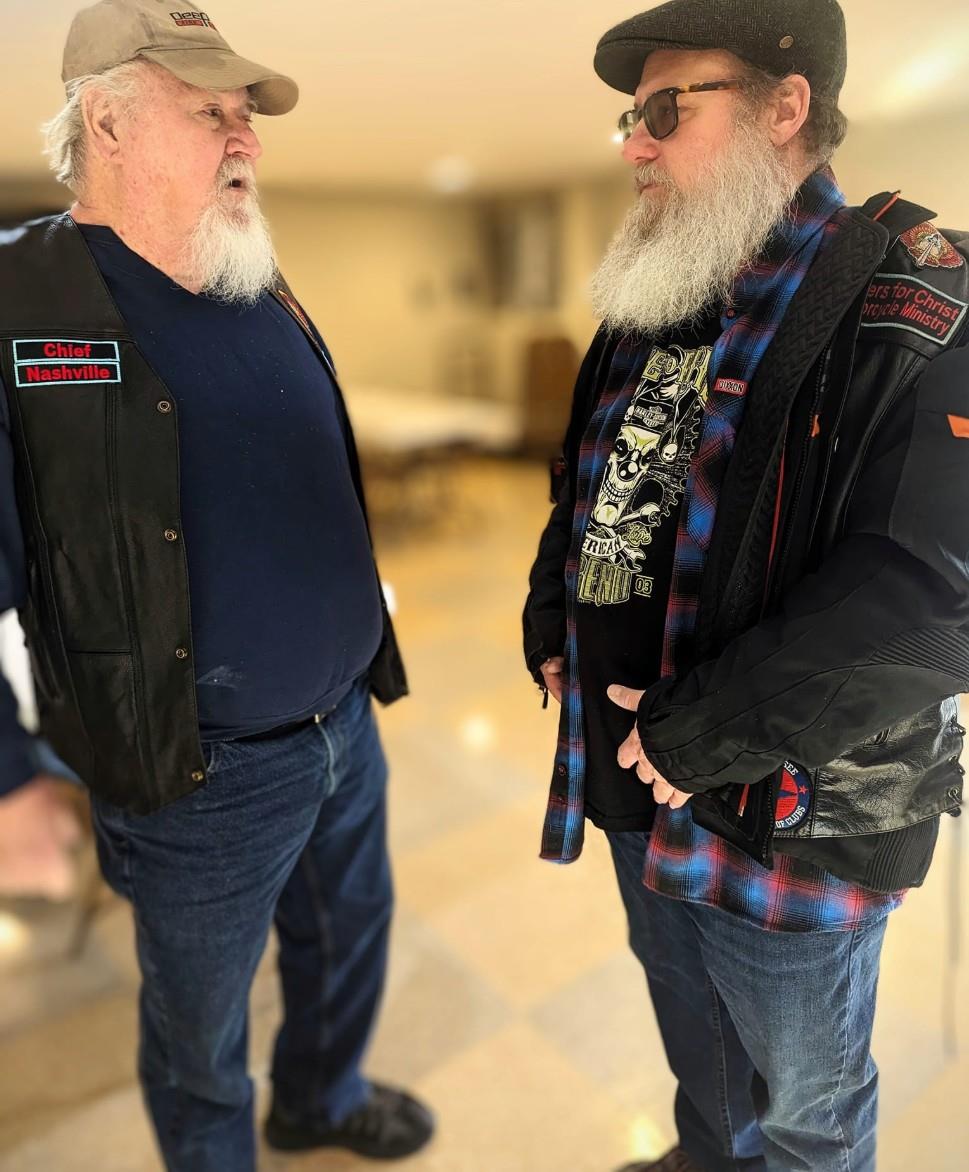 Two men with long beards talk with each other indoors, dressed in casual jackets and jeans.