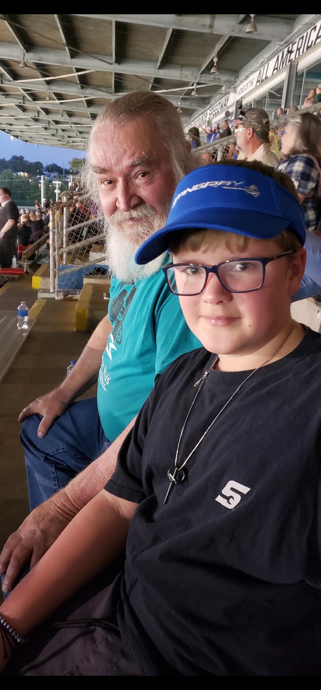 A boy in glasses sits with an older man at a lively sports event, both enjoying it.