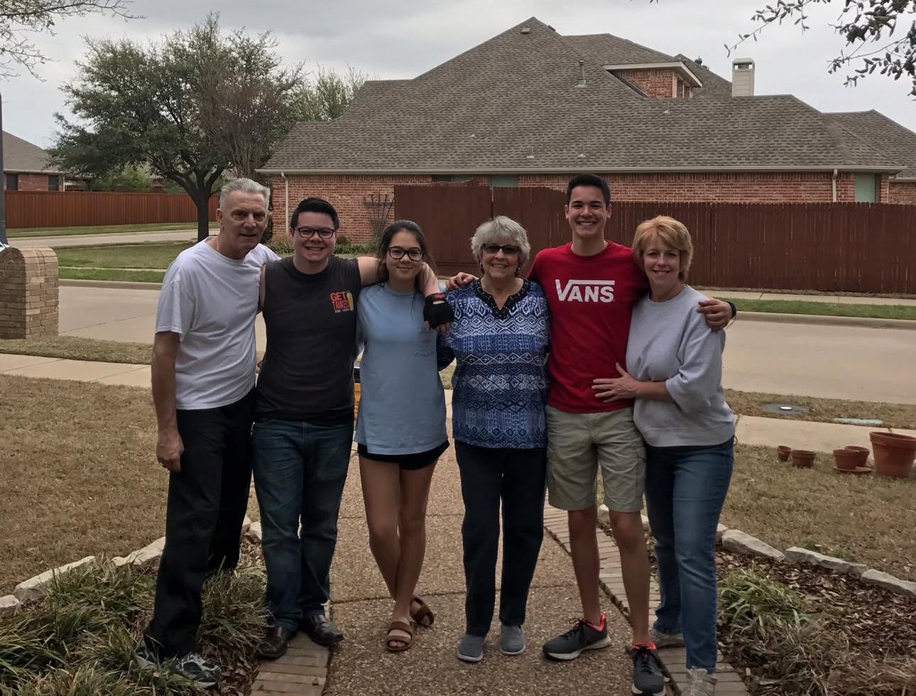 Group of six family members standing together in a front yard outside a house on a cloudy day.