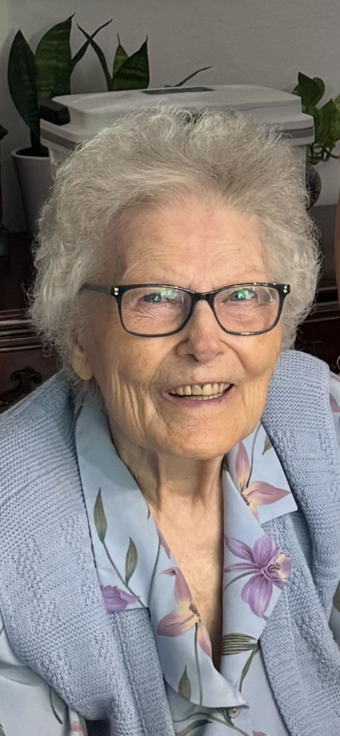 An elderly woman with curly white hair and glasses smiles happily, radiating joy in her cozy home.