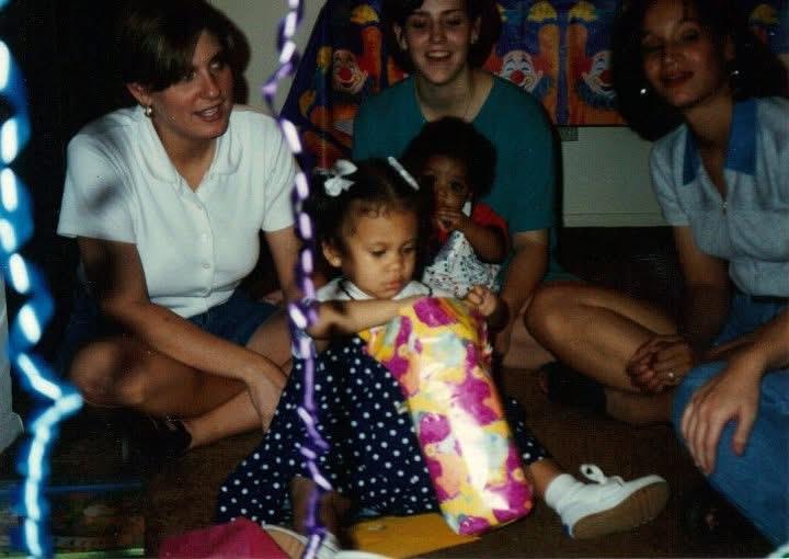 A young girl with a bow in her hair is excitedly unwrapping a gift surrounded by family members.