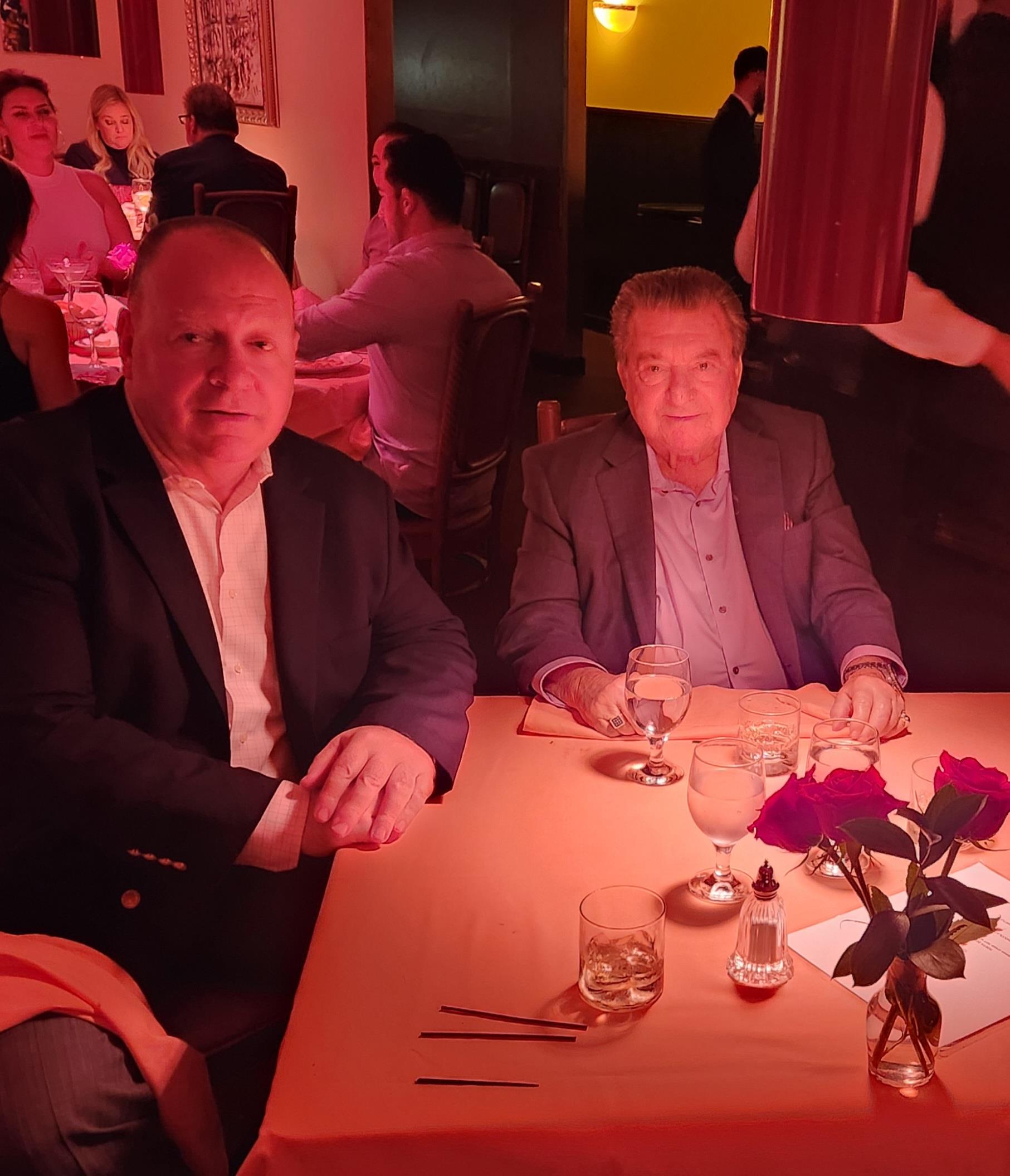 Two men are seated at a table in a dimly lit restaurant, enjoying dinner with drinks.