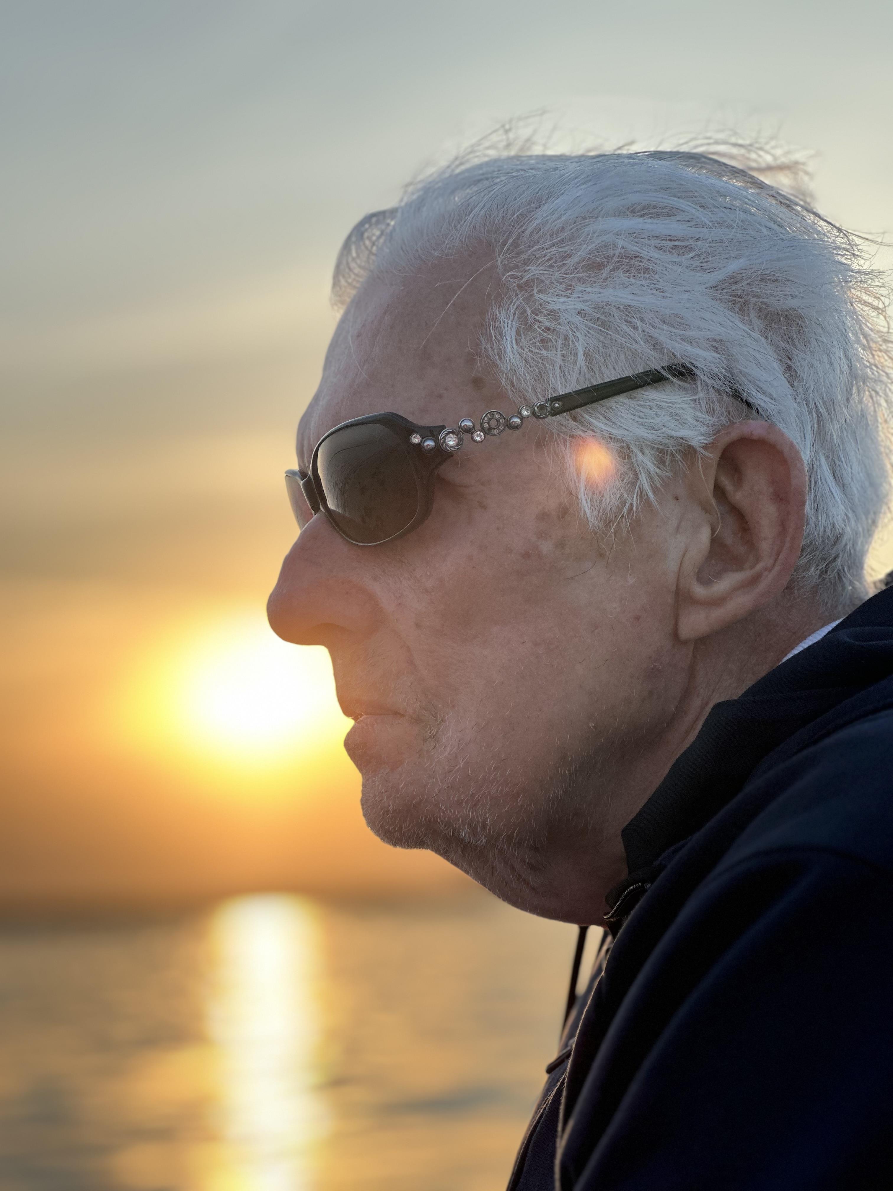 Senior man watches the sunset with a serene expression, wearing dark sunglasses near the water.