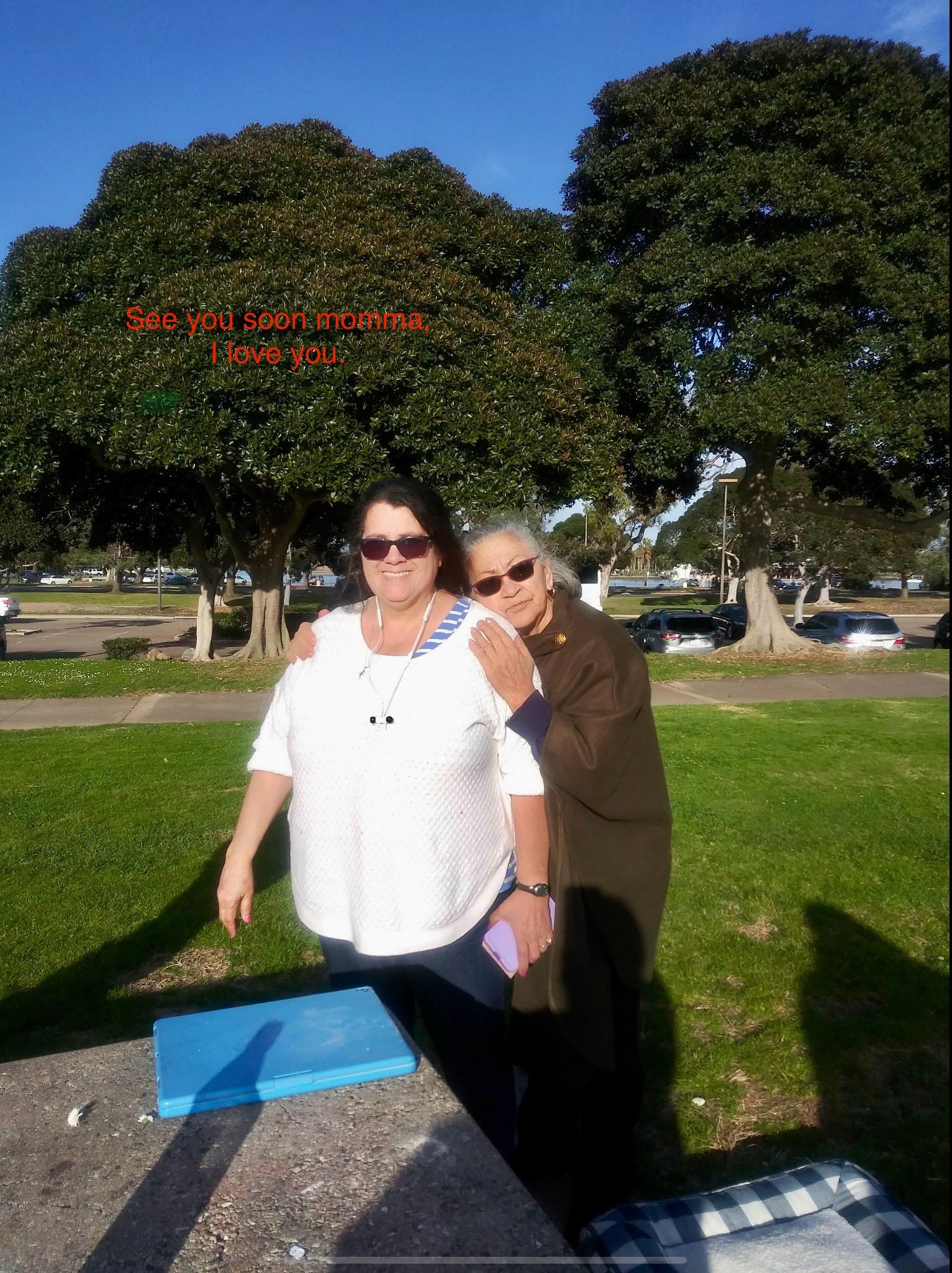 Two women stand together outside, smiling and enjoying the pleasant weather in a park setting.