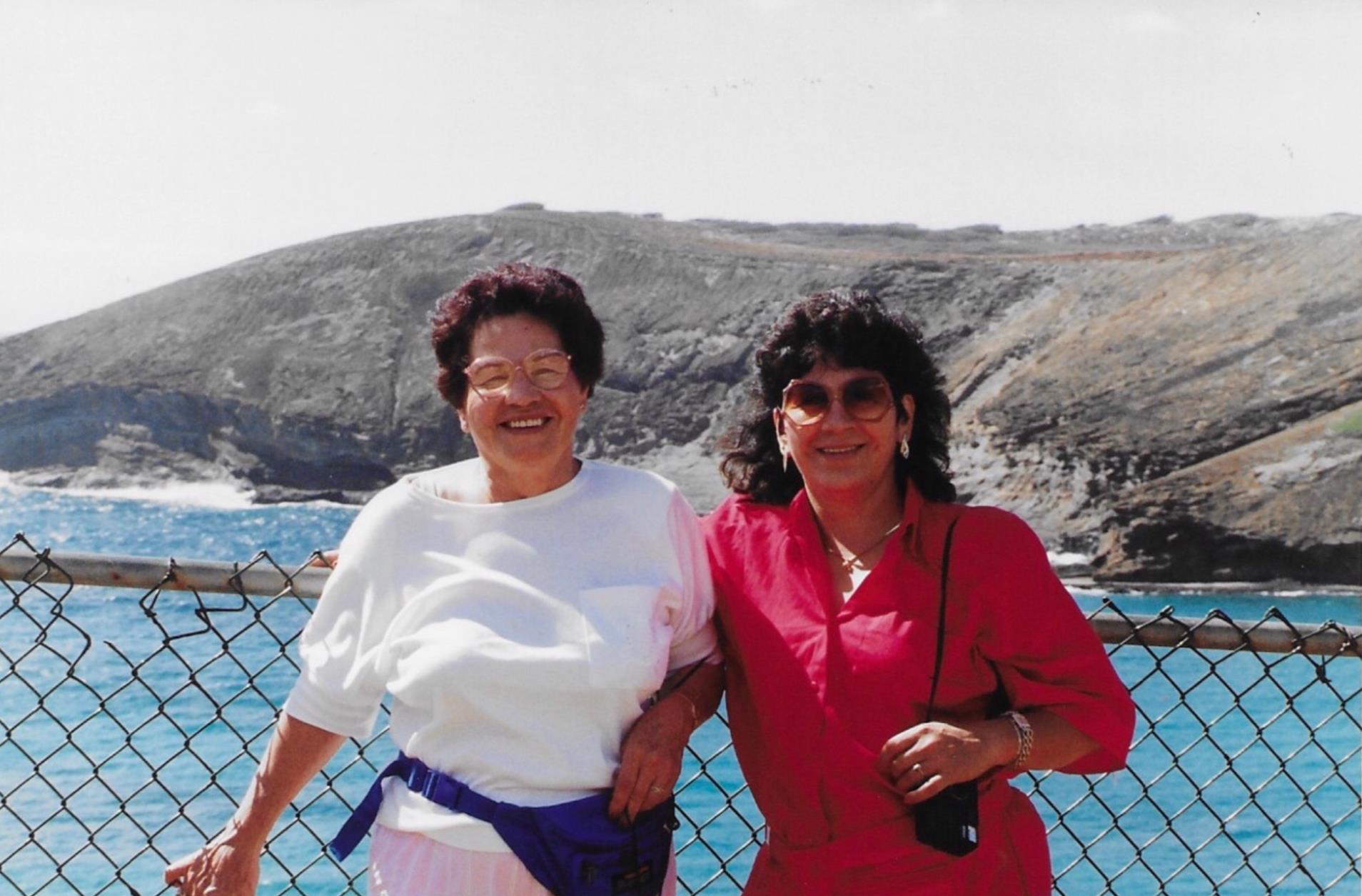 Two women stand together by the shore, smiling against a backdrop of mountains and ocean waves.