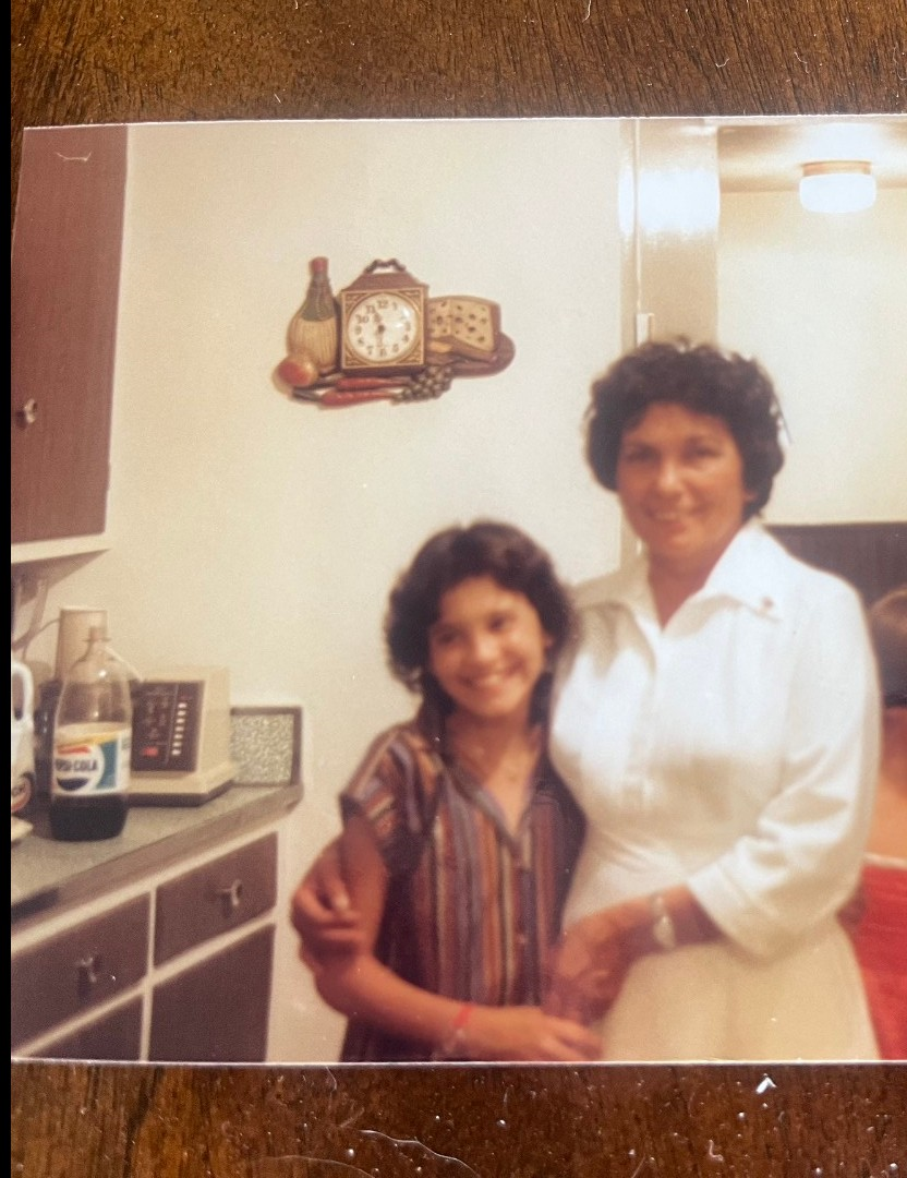 A woman and her daughter smile together in a warmly lit kitchen from the 1970s, radiating happiness.