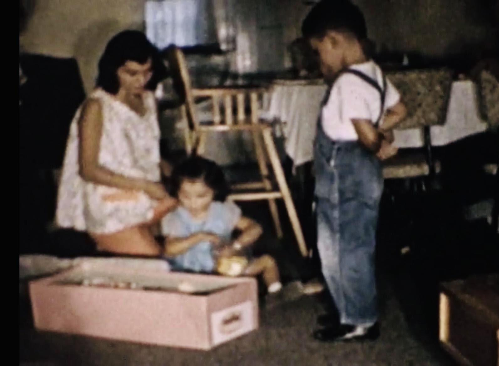 A mother helps her children enjoy a playful moment with a pink box in their living room.