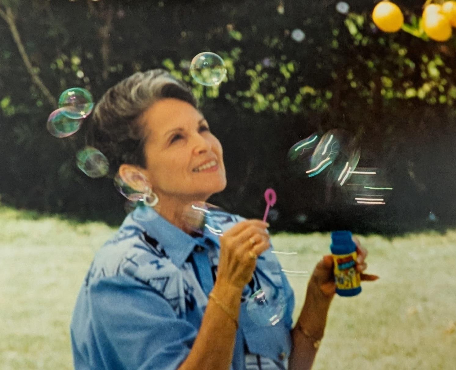 A woman smiles joyfully while blowing bubbles on a bright, sunny day in a garden.