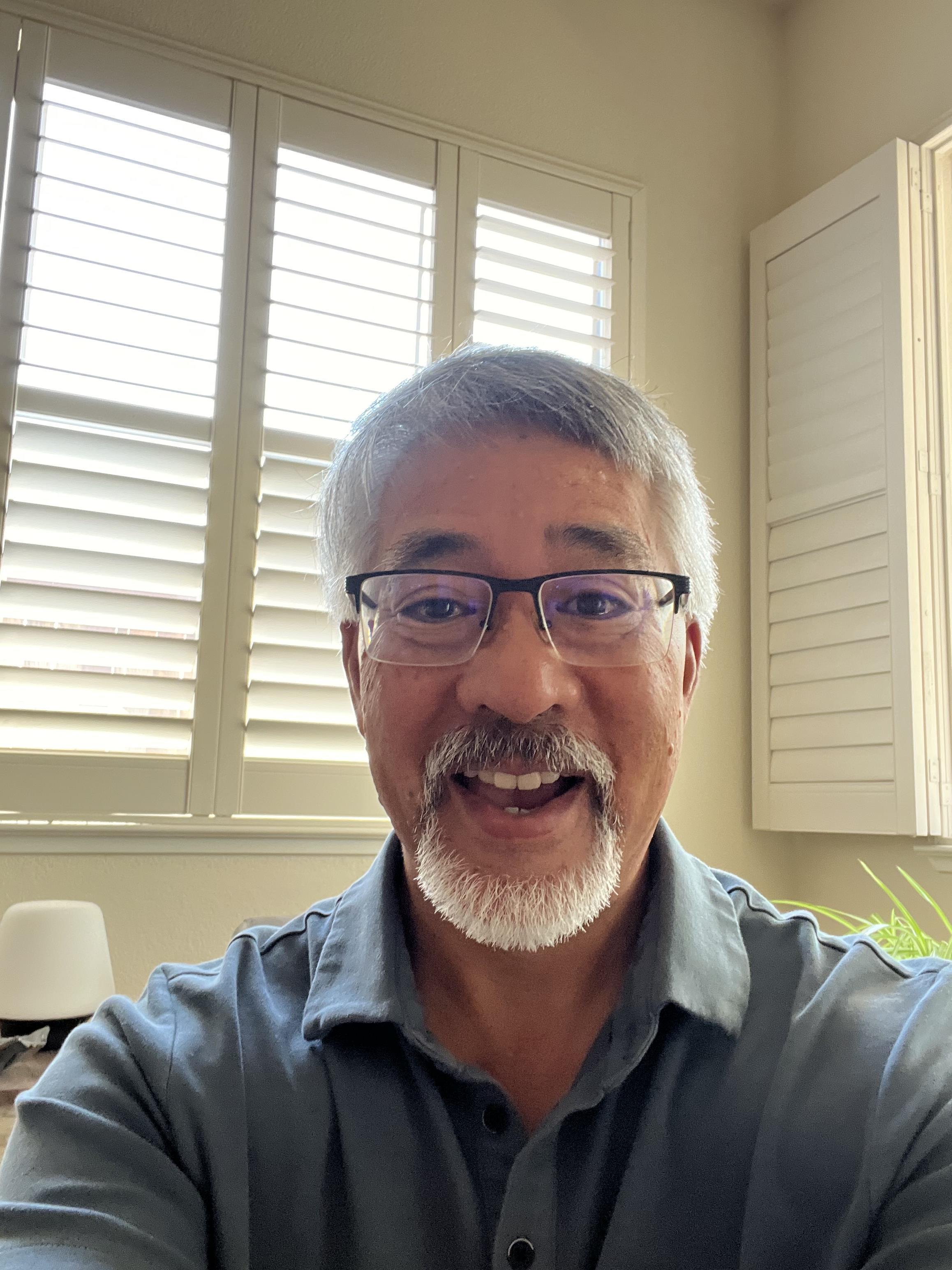 A joyful older man is smiling at the camera in a well-lit room with greenery.