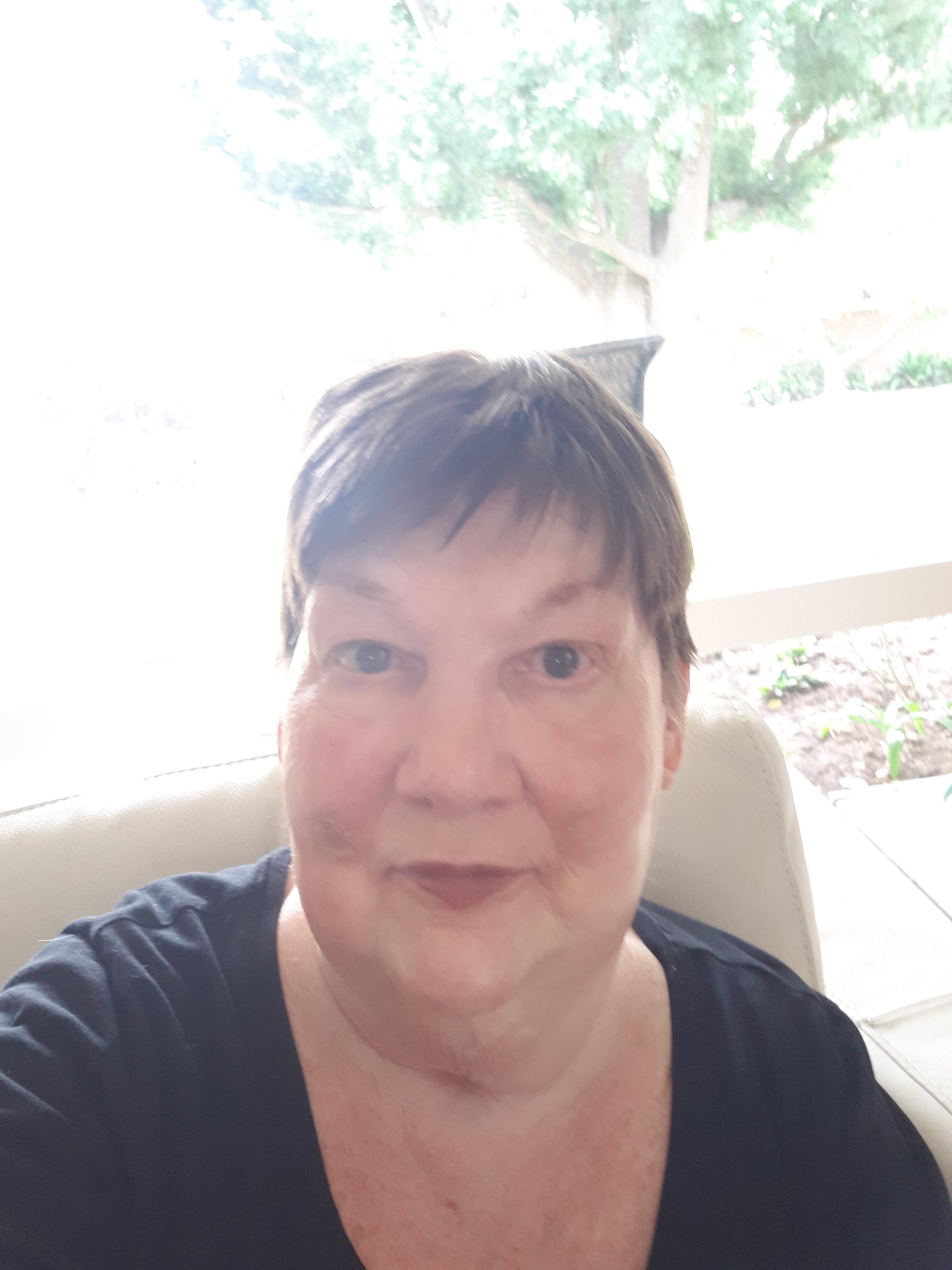 A woman with short hair smiles warmly while sitting indoors, basking in soft daylight.