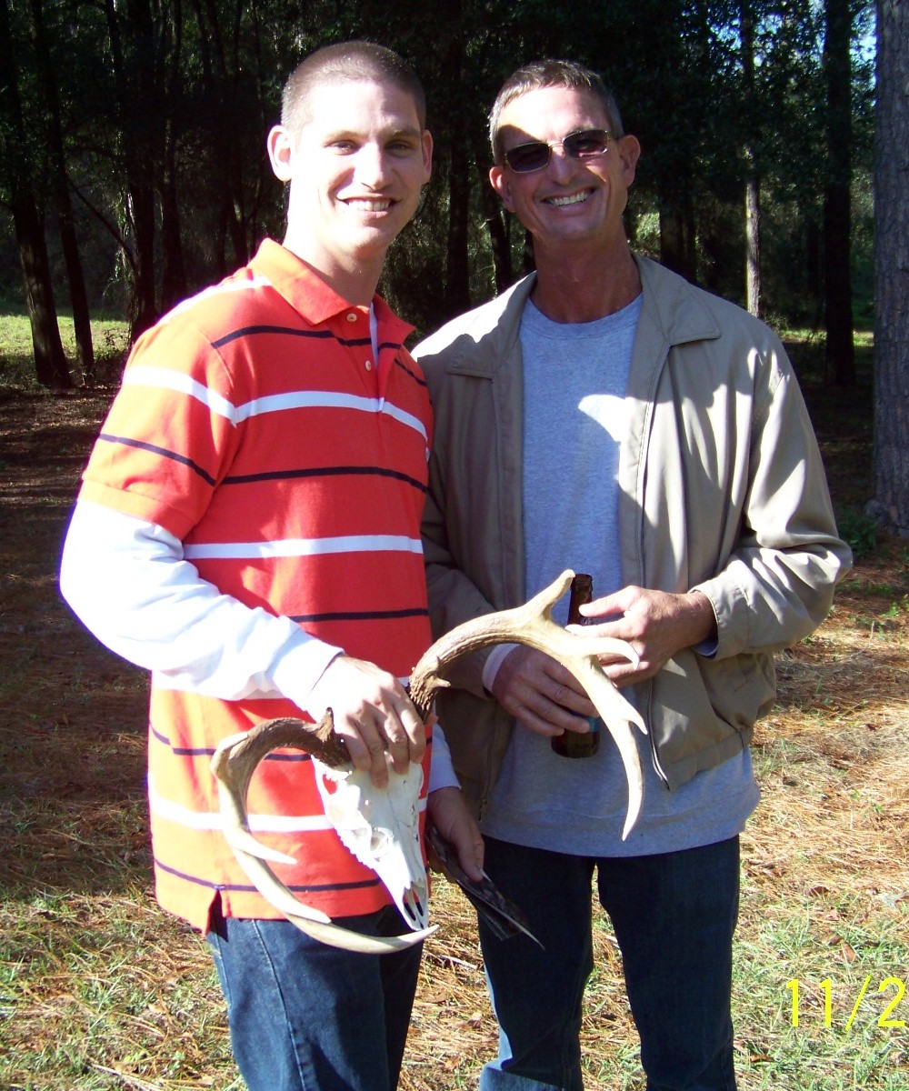 Two men smile as they hold deer antlers, enjoying a sunny day in a wooded area during autumn.