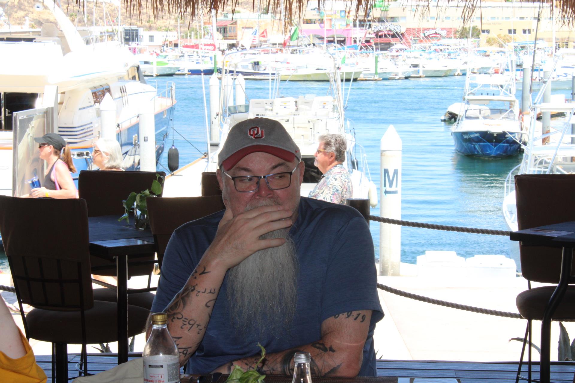 A bearded man with tattoos enjoys his meal at a table by the marina as boats float past.