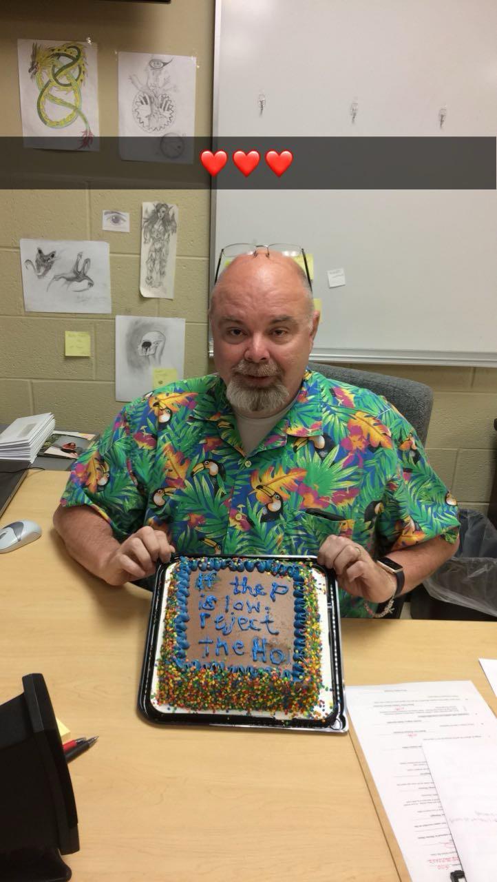 A cheerful teacher in a colorful shirt holds a birthday cake as students celebrate.
