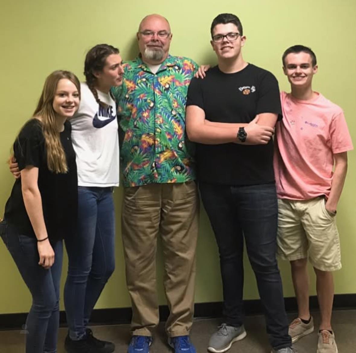 Five friends pose playfully with a teacher during a casual gathering in a school hallway.