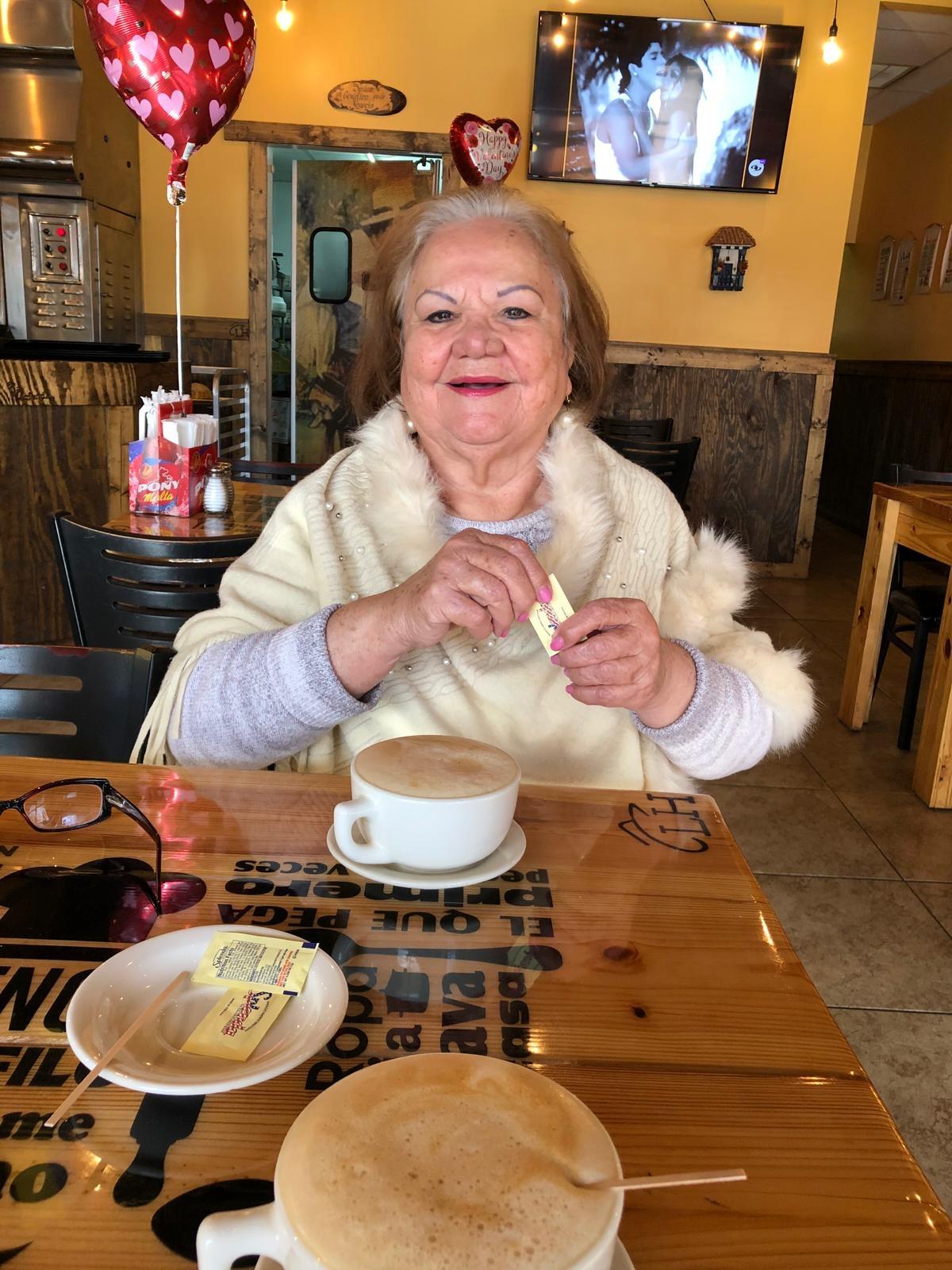 A cheerful older woman adds sweetener to her coffee while seated at a wooden table.