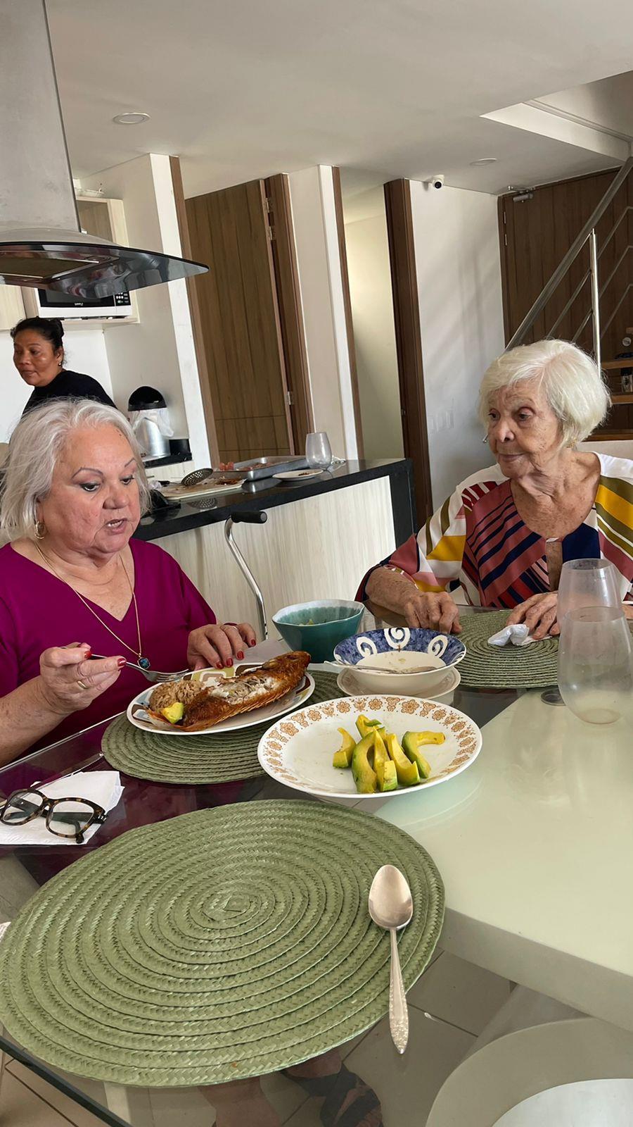 Two elderly women share a meal of fruits and pancakes in a bright kitchen while chatting.