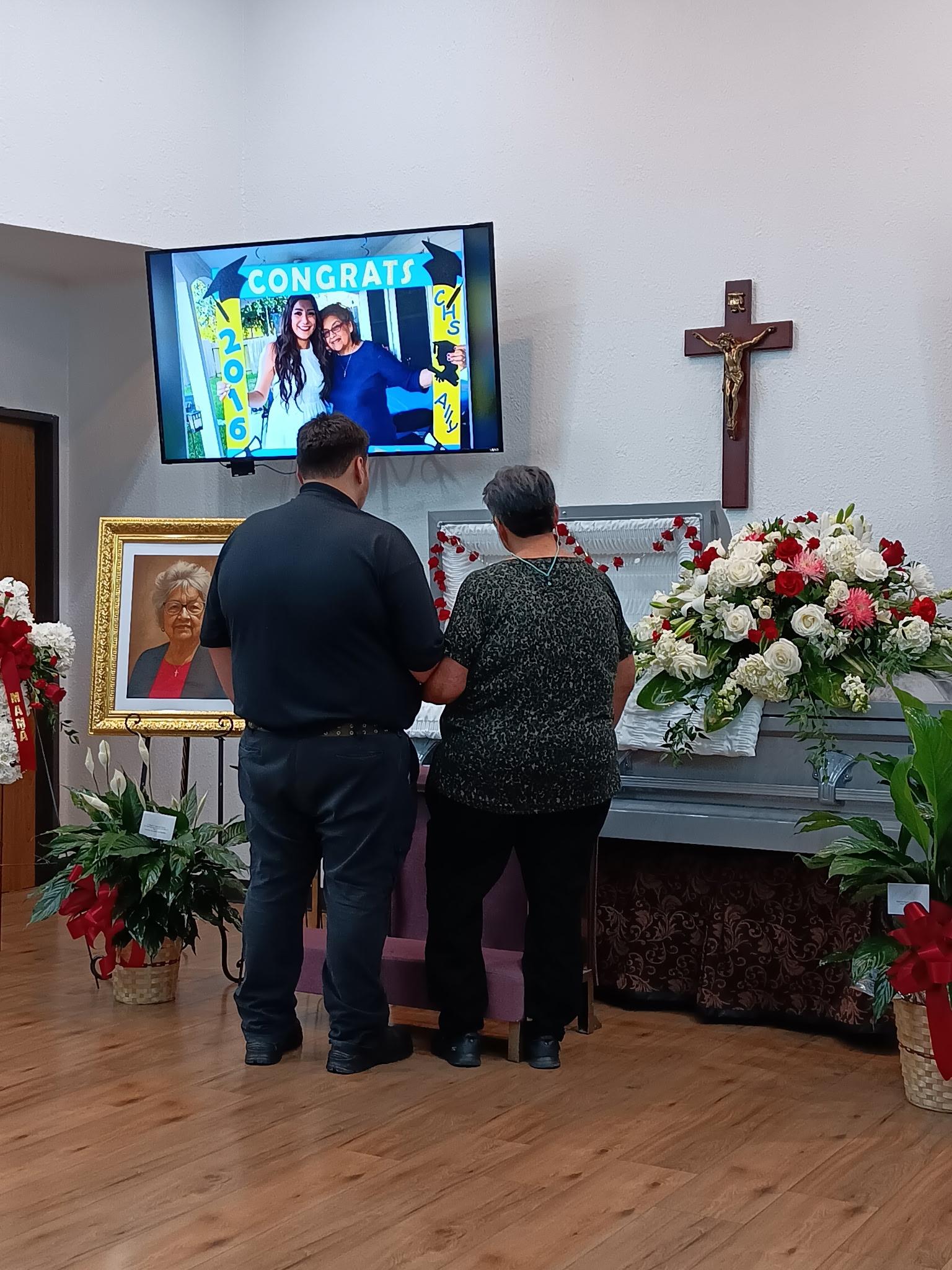 Two people stand at a memorial, surrounded by flowers and a congratulatory screen.