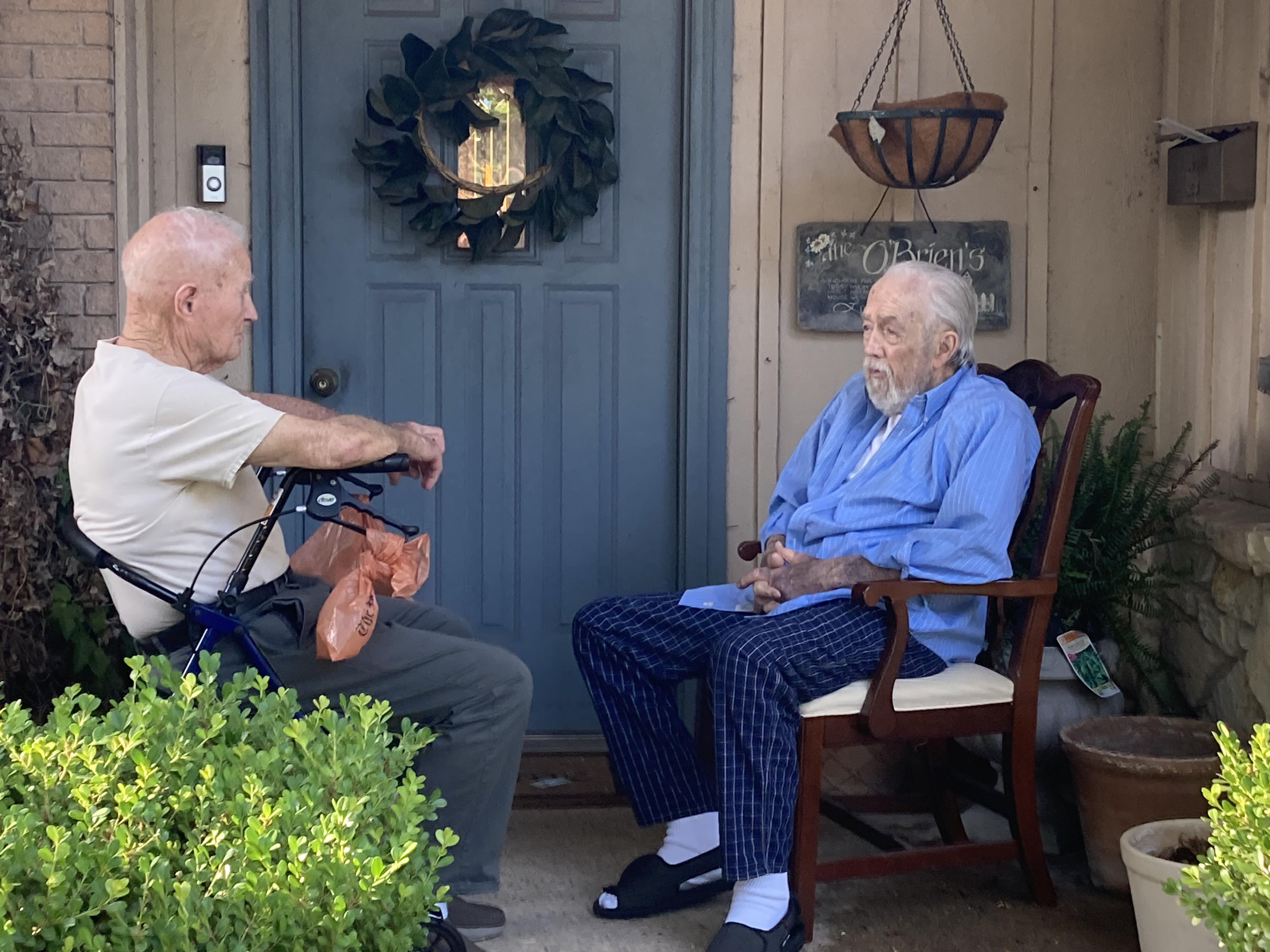 Two elderly men sit on a porch, engaged in conversation while enjoying a warm sunny afternoon.