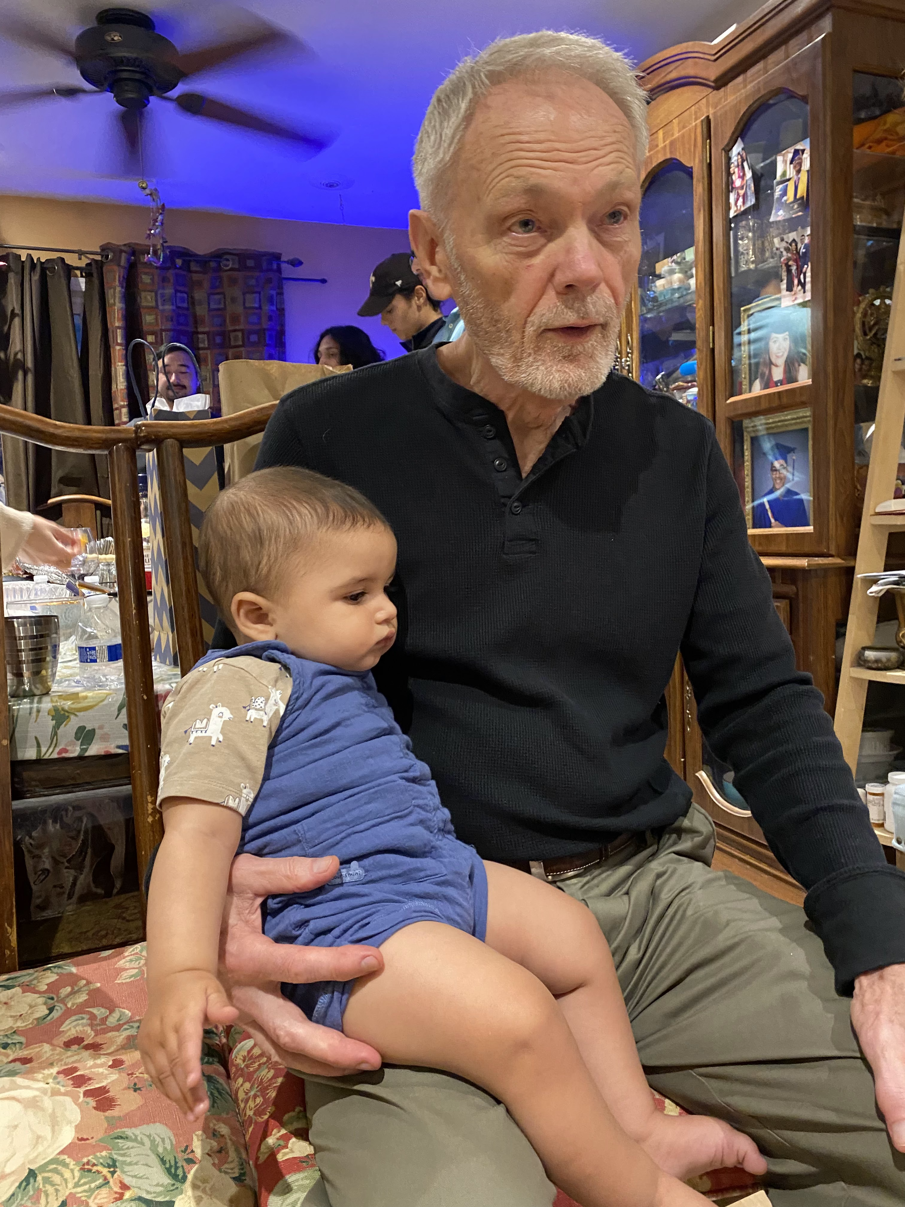 An elderly man sits with a baby on his lap in a cozy indoor setting filled with family.