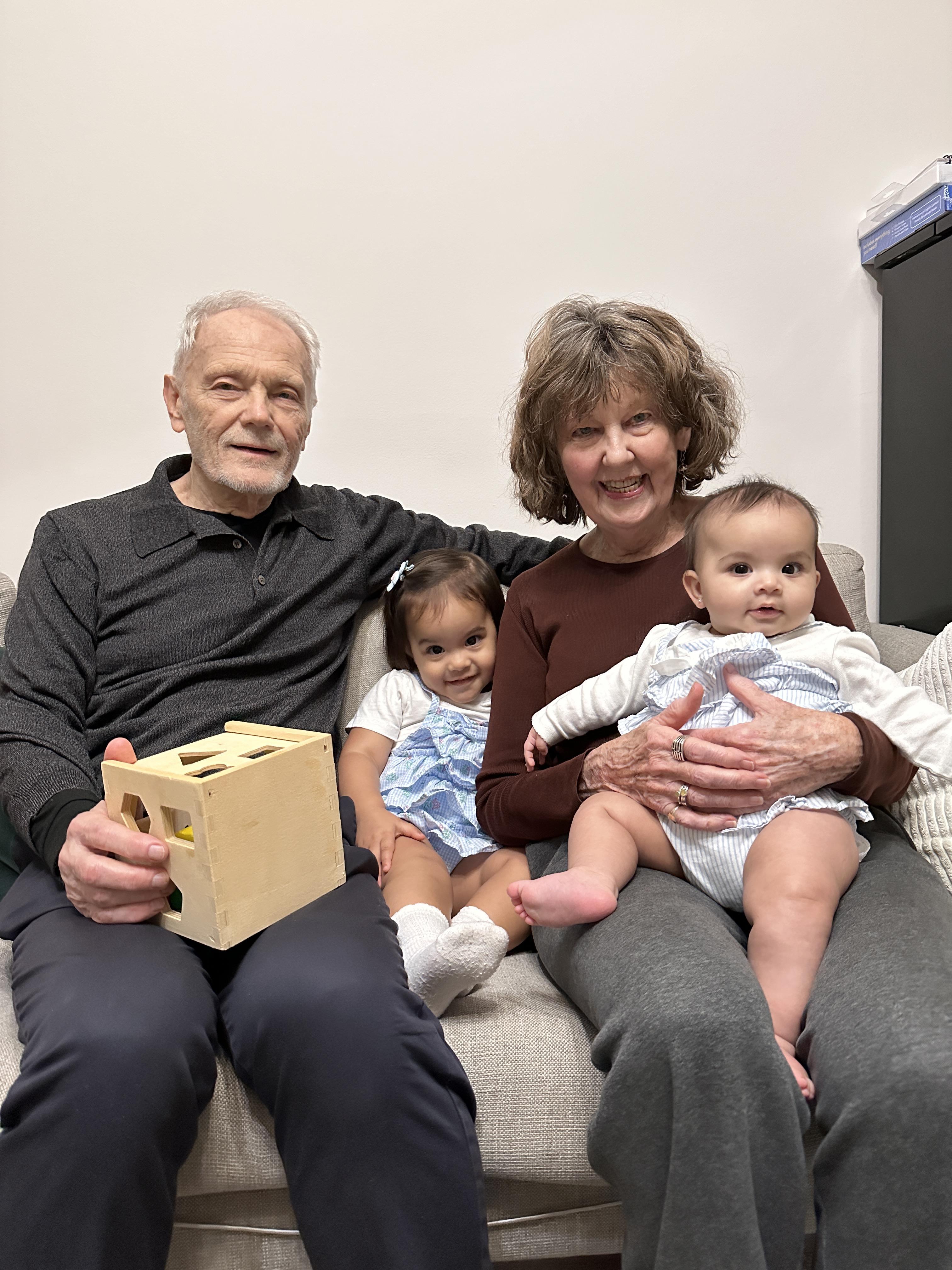 Grandparents sit on a sofa with their two young grandchildren, sharing joyful moments together.