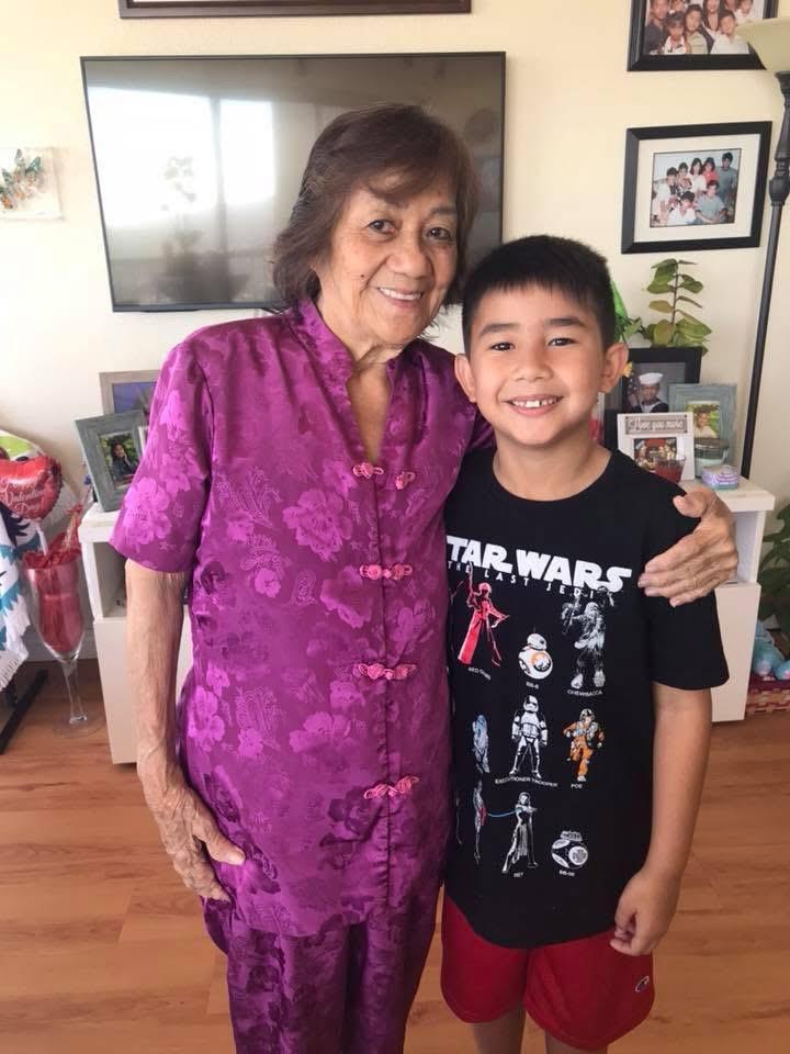 A young boy smiles next to an elderly woman wearing a purple dress in a home environment.