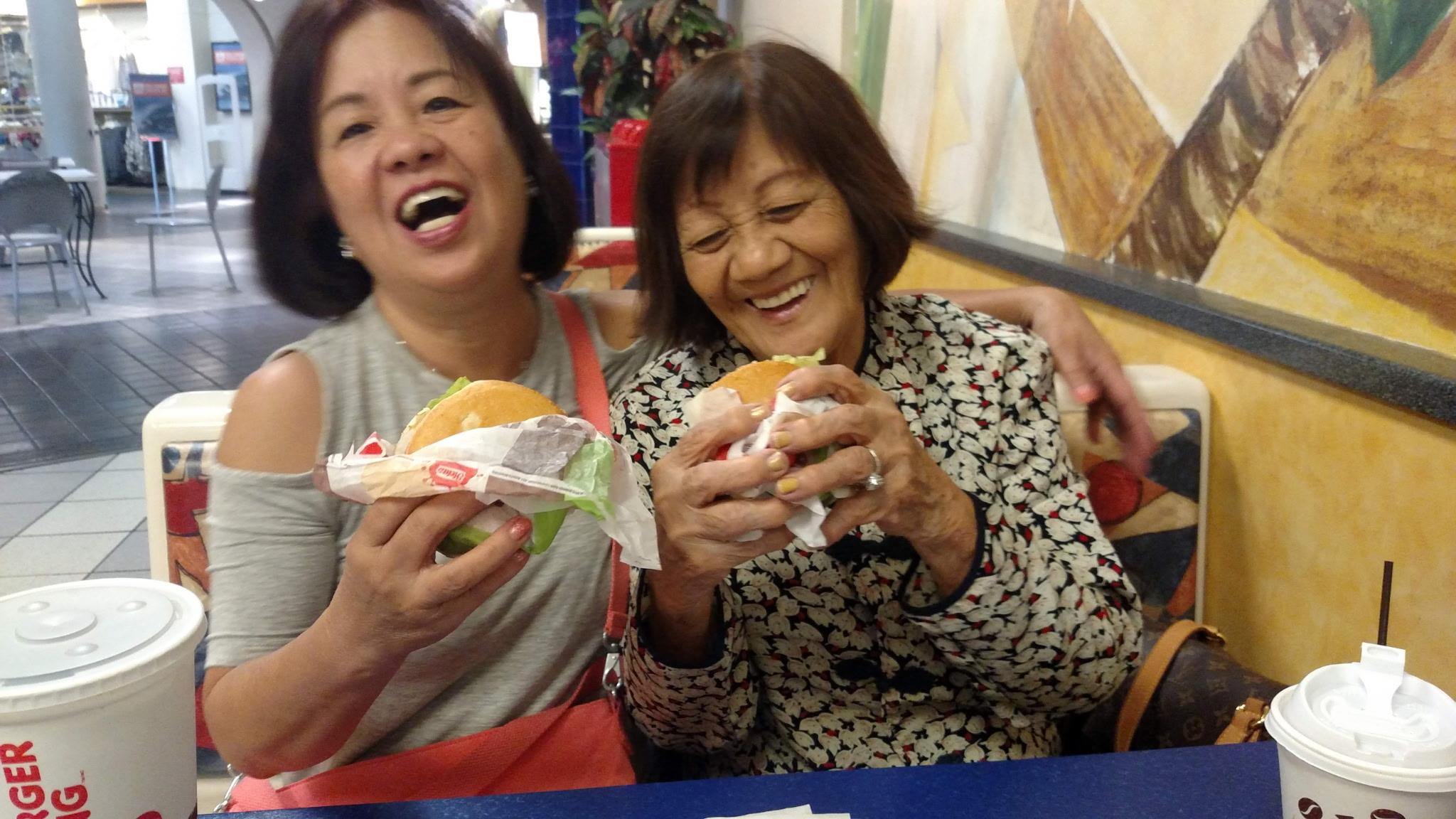 Two joyful women laugh and share a moment while holding burgers in a crowded restaurant.