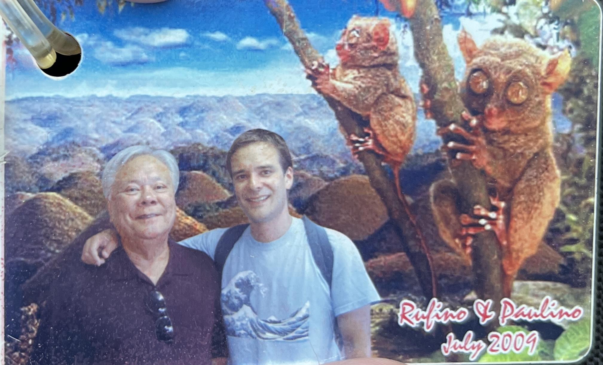Two men stand together in front of a colorful mural depicting trees and fantasy creatures.