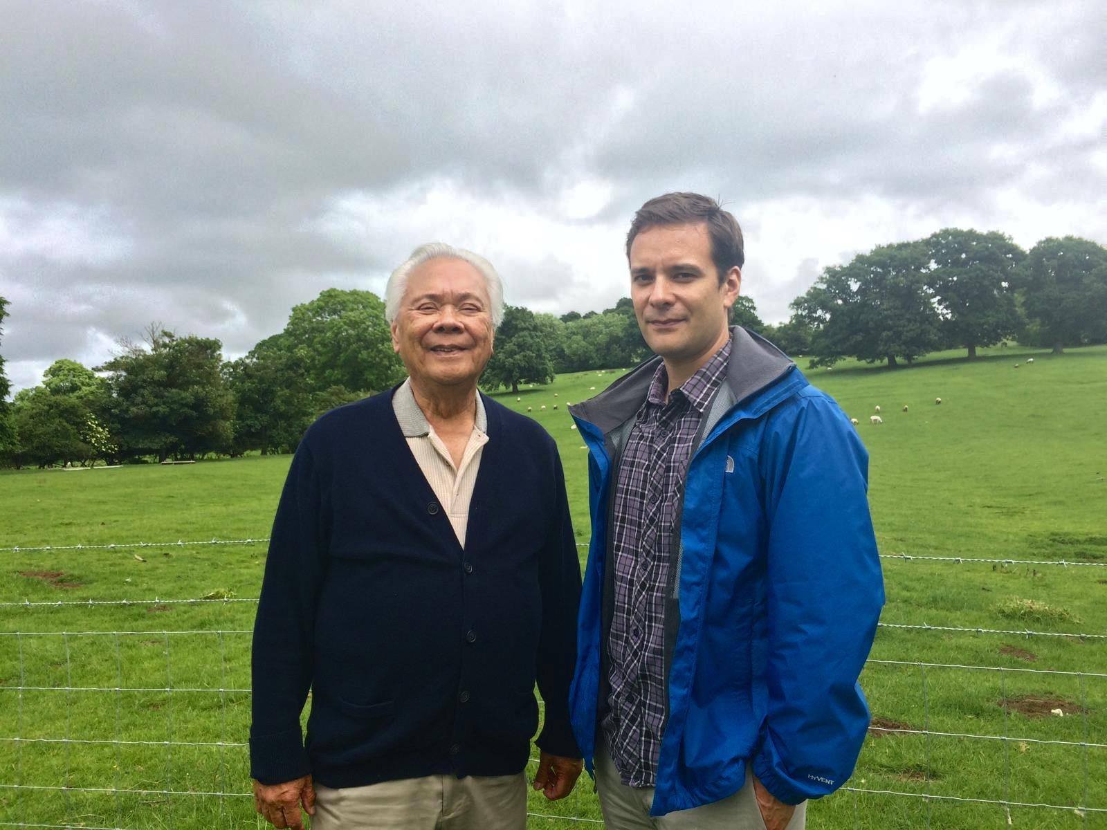 Two men posing in a lush green field, surrounded by trees, under an overcast sky.