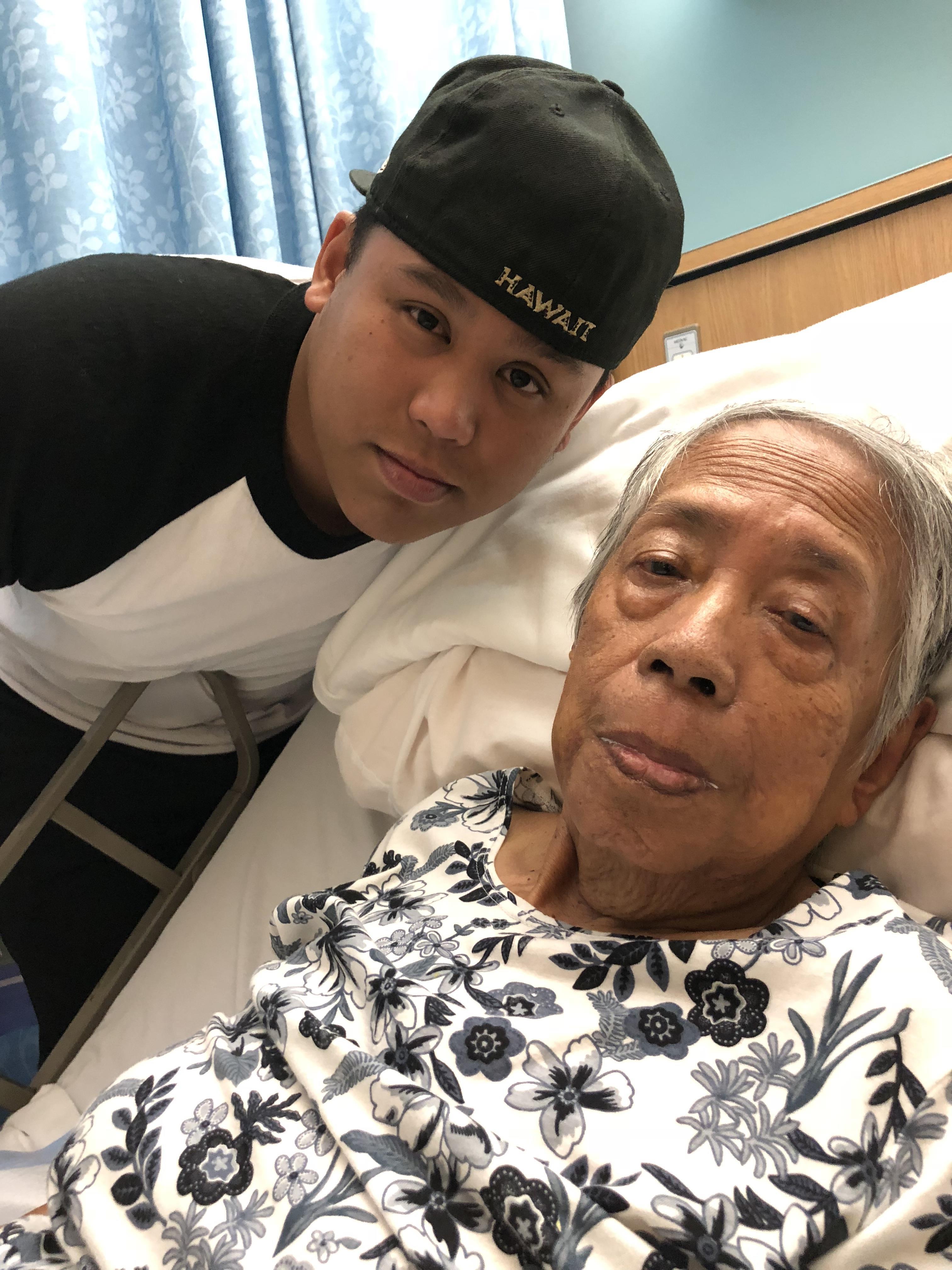 A young man leans close to an elderly woman in a hospital room, both looking at the camera.