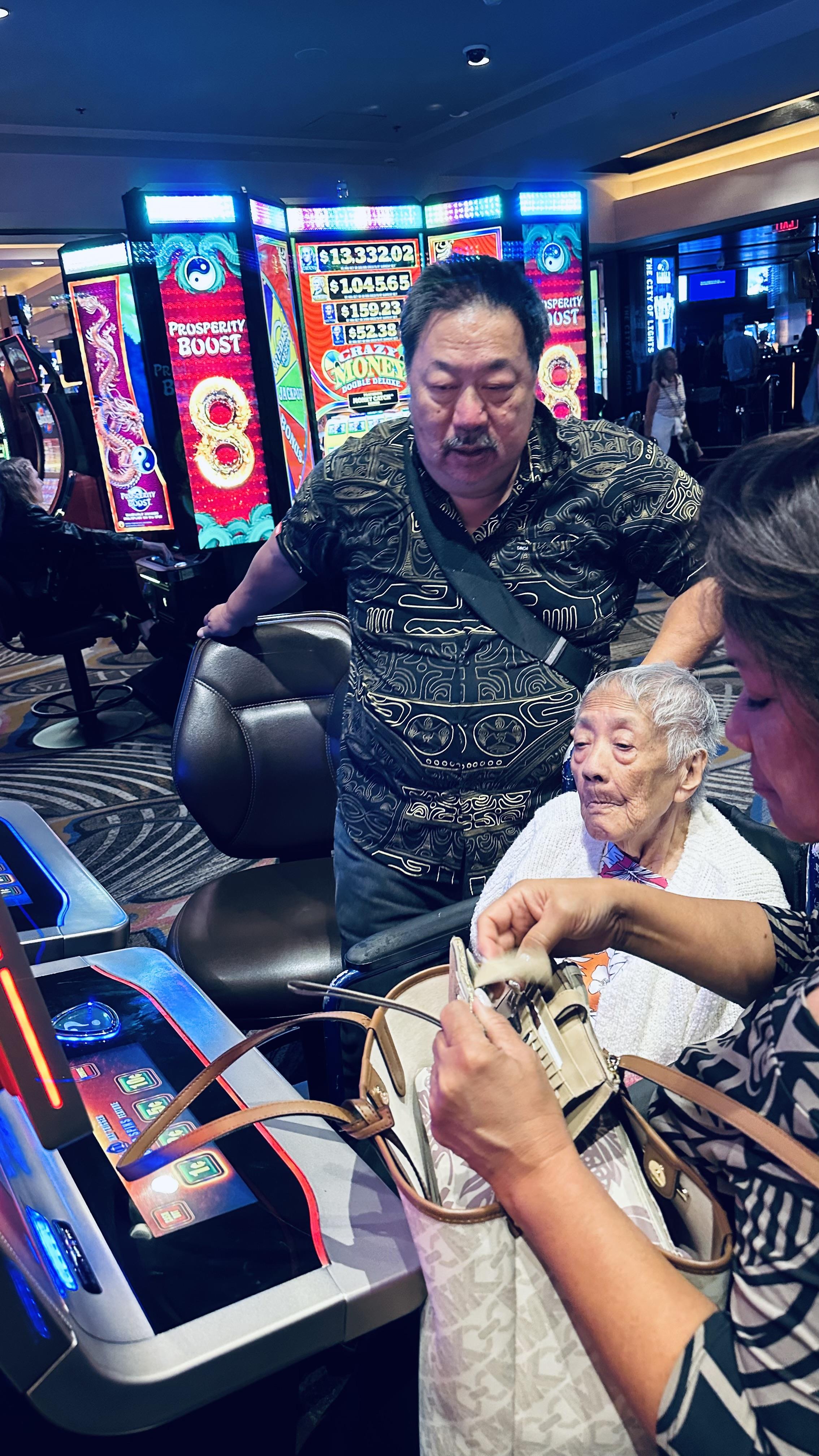 Two family members assist an elderly woman at a casino slot machine during the night.