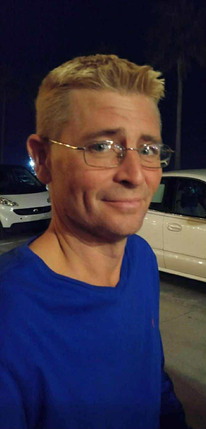 A man with glasses smiles warmly, dressed in a blue shirt, surrounded by parked vehicles at night.