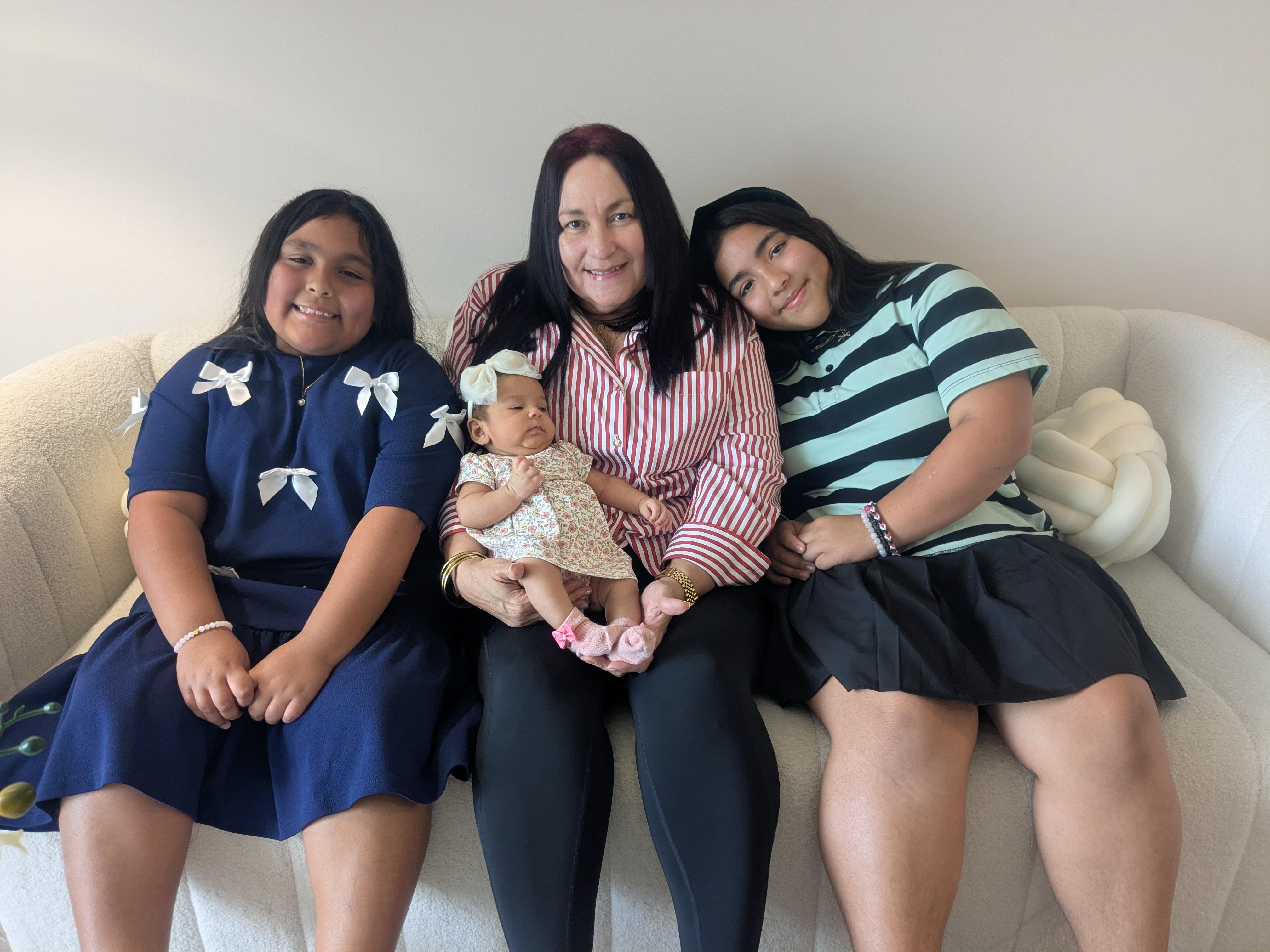 A mother sits with her two daughters, holding a baby, all smiling happily together in a living room.