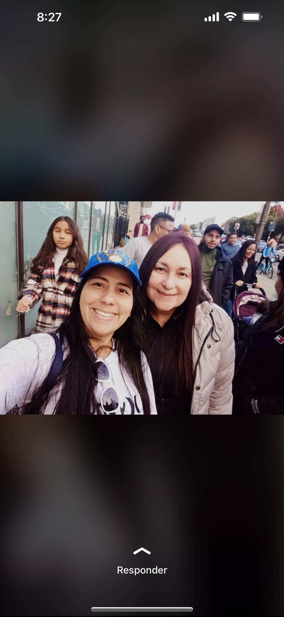 Two women pose happily for a selfie while a crowd walks by, enjoying a vibrant community event.